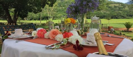 Une table à manger installée à l’extérieur, recouverte de nappes blanches, agrémentée de centres de table floraux et d’une bouteille de champagne.