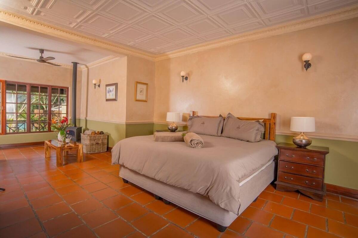 A spacious bedroom with a large bed, a nightstand, a small table with a vase, and a window with a view of greenery.