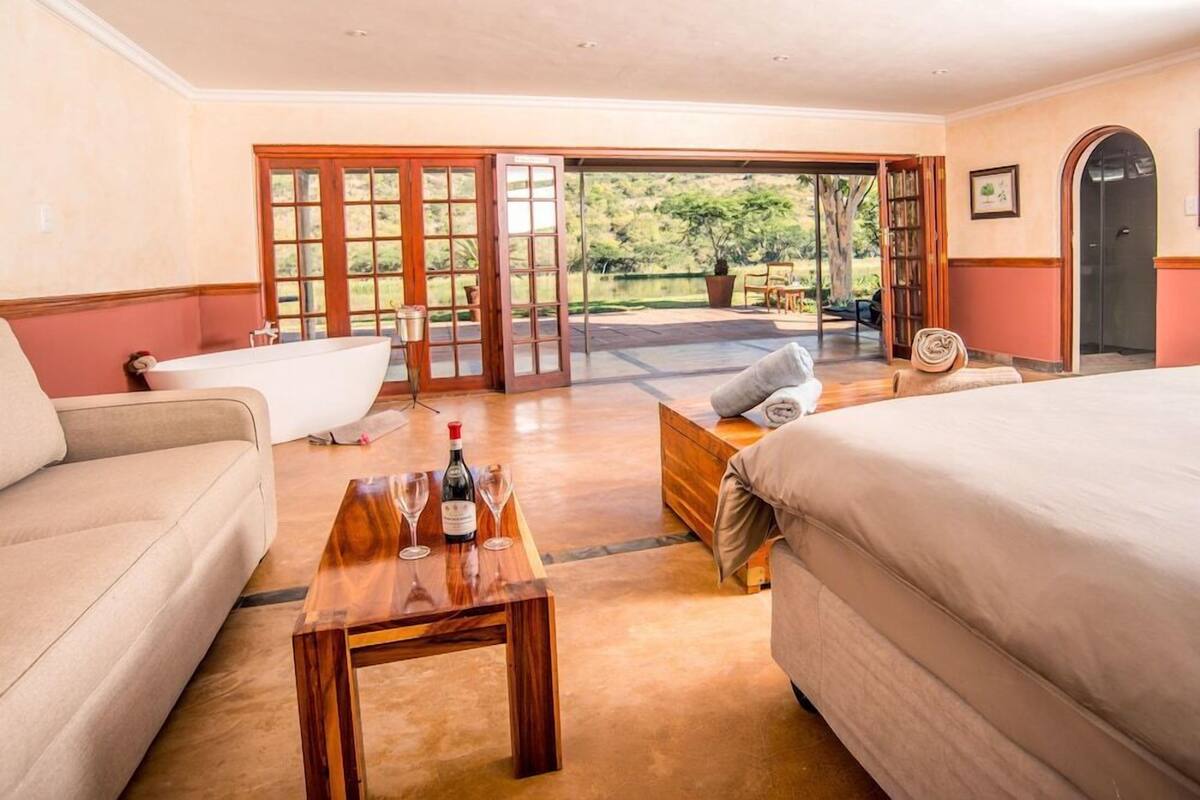 A room with two beds, a sofa, a wooden table with wine glasses, and a view of a garden through glass doors.