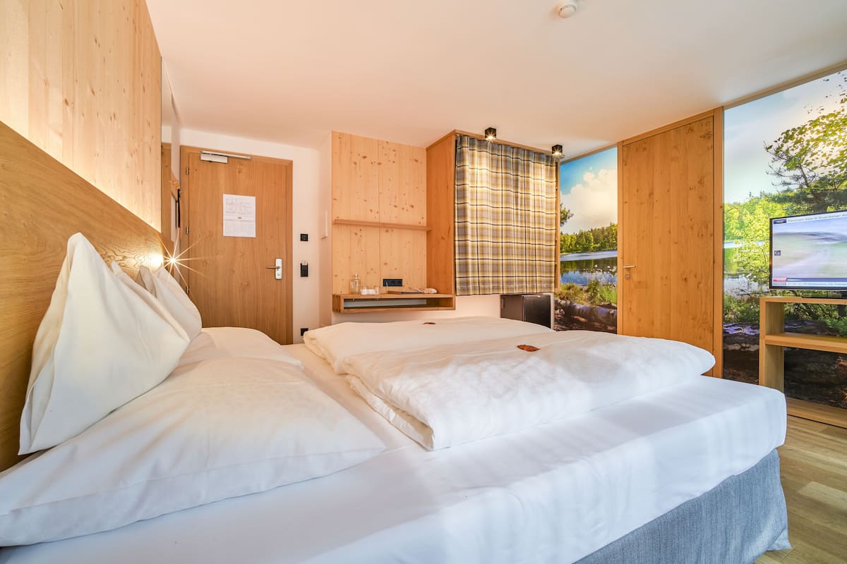 Une chambre avec un grand lit, des murs en bois, un petit bureau et une fenêtre donnant sur des arbres.