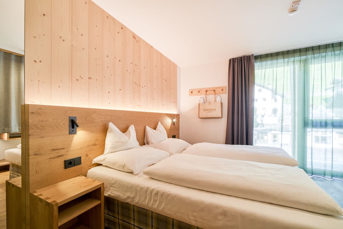 Une chambre d’hôtel avec une tête de lit en bois, un lit avec une literie blanche, une table de chevet et une vue sur des immeubles à travers une fenêtre avec des rideaux.