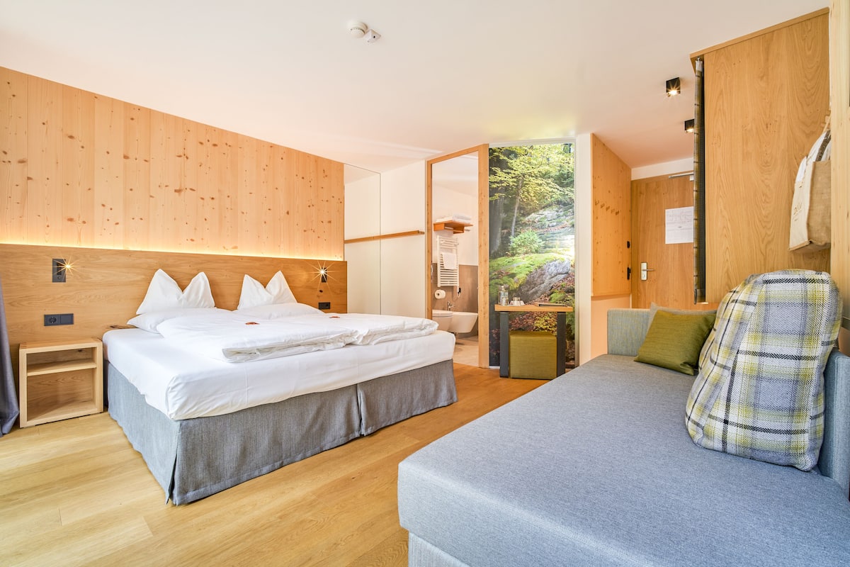 Une chambre d’hôtel moderne avec un mur en bois, un grand lit, un canapé et une vue sur une forêt, visible à travers une porte vitrée.