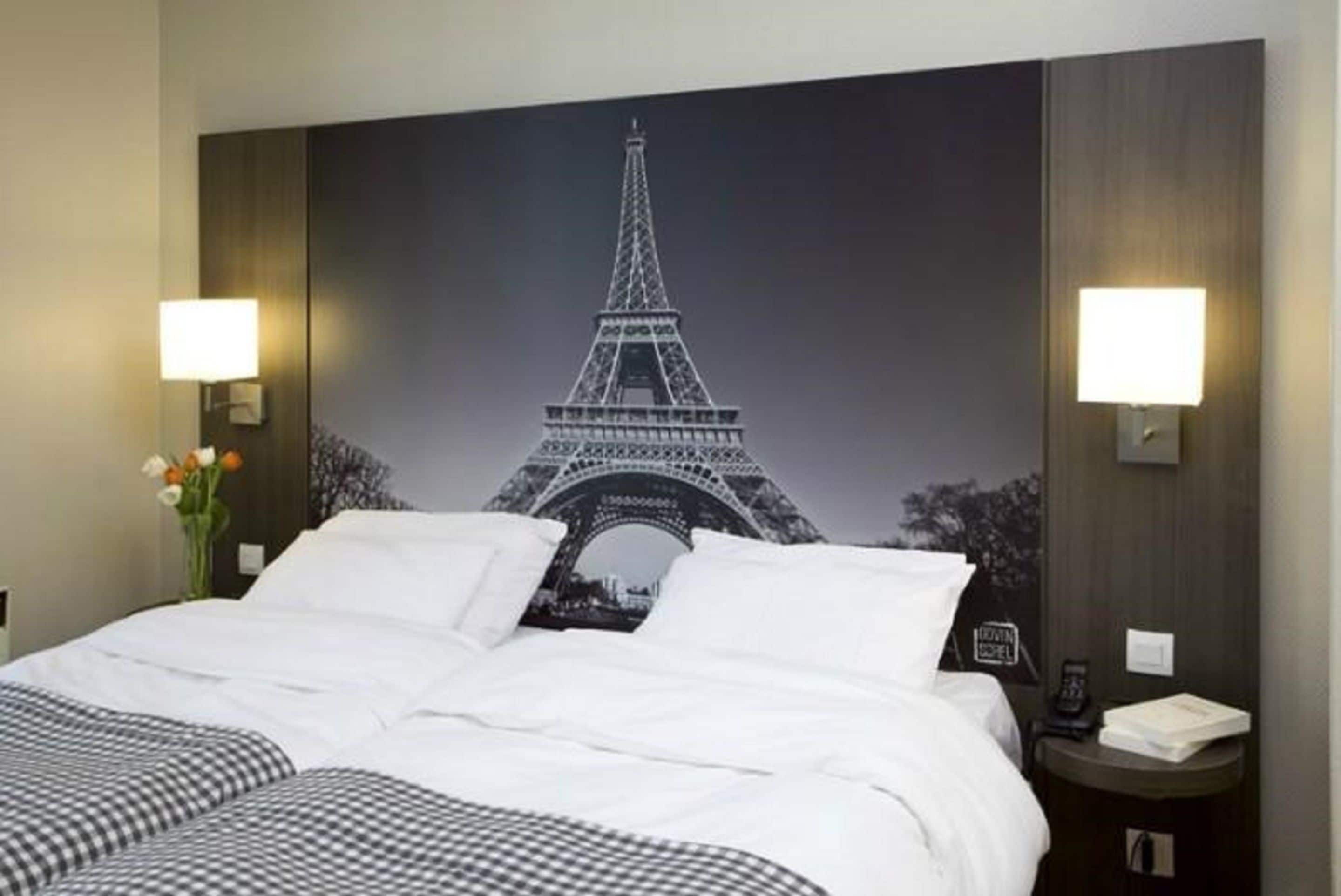 A hotel room with a large wall mural of the Eiffel Tower, a bed with checkered bedding, two bedside lamps, and a nightstand with a phone and a book.