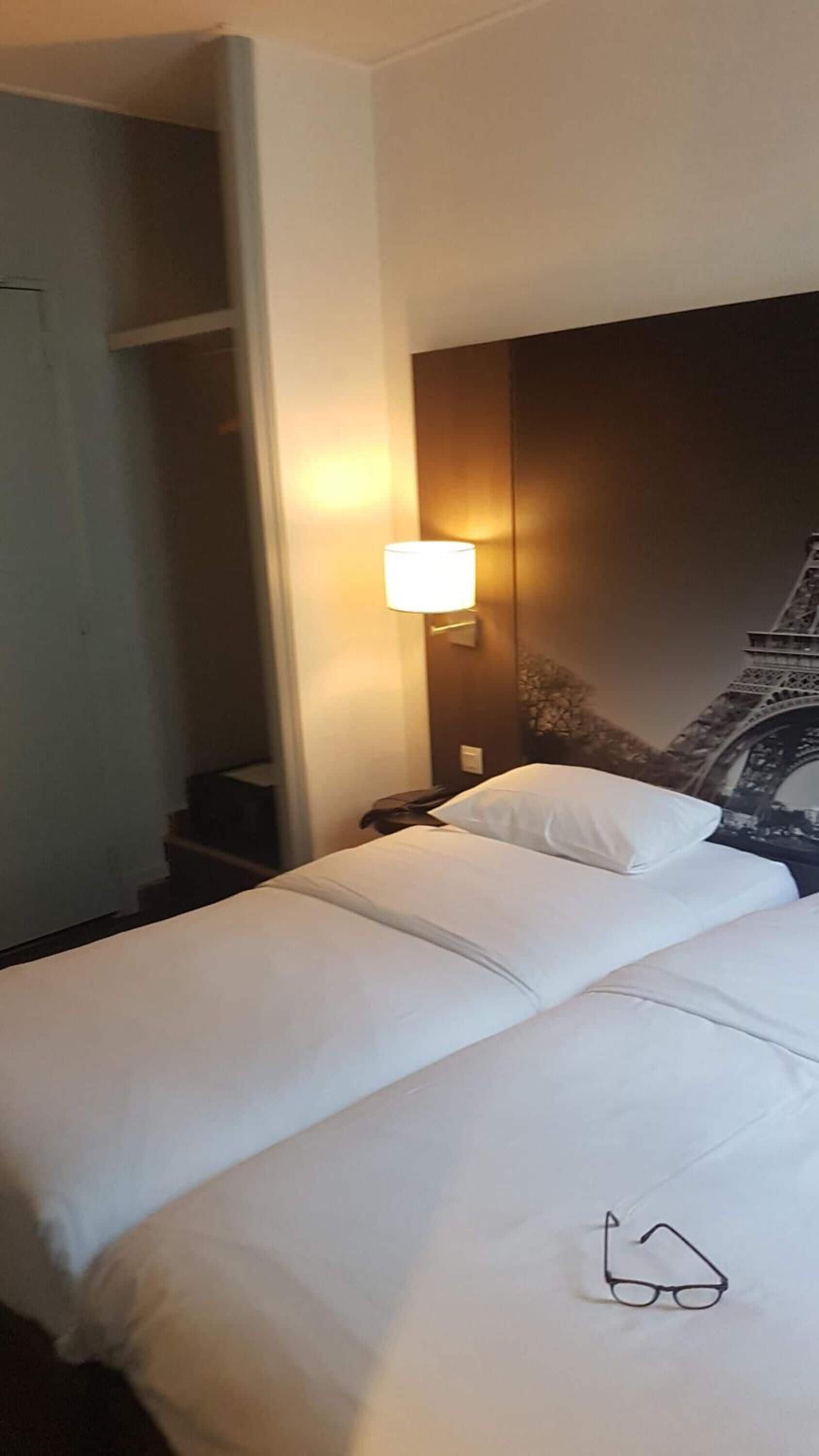 A hotel room with a bed, a nightstand, a lamp, and a wall-mounted picture of the Eiffel Tower.