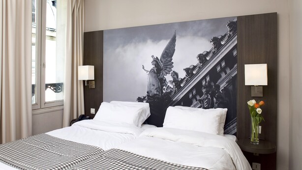 A hotel room with a large bed, two bedside tables with lamps, a large framed picture on the wall, and a vase with flowers on the nightstand. - Hôtel Victoria Paris (Paris)