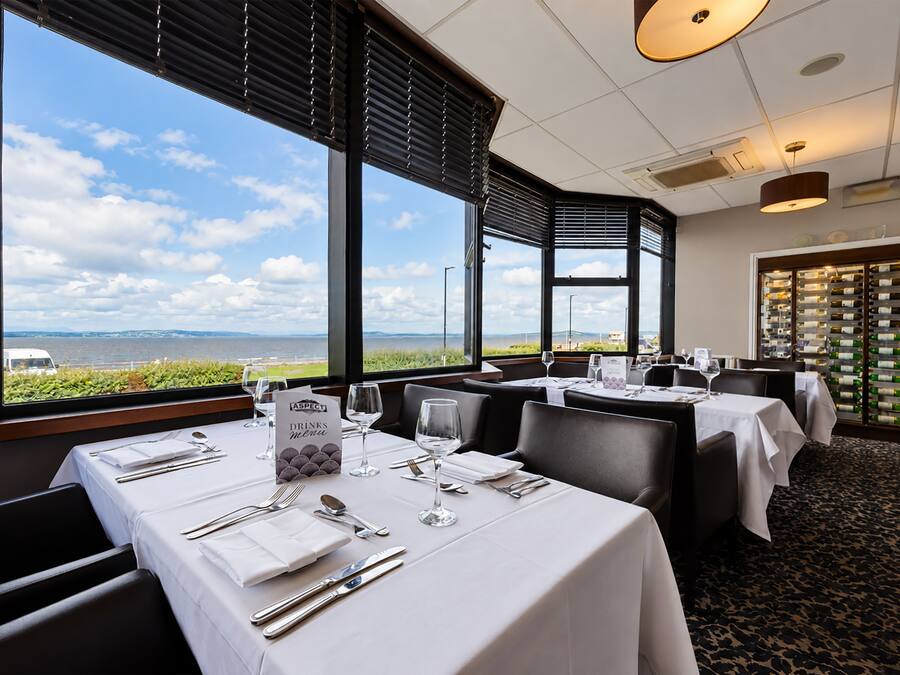 Un restaurante con mesas dispuestas para comer, amplias ventanas con vistas al mar y una estantería con botellas de vino al fondo.