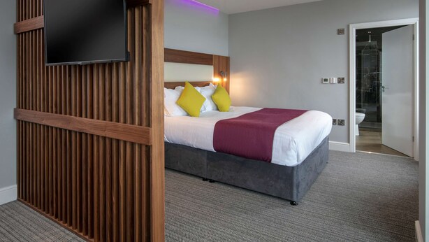 Una habitación de hotel moderna con una cama grande, un televisor de pantalla plana montado sobre un panel de madera y un baño visible a través de una puerta abierta.