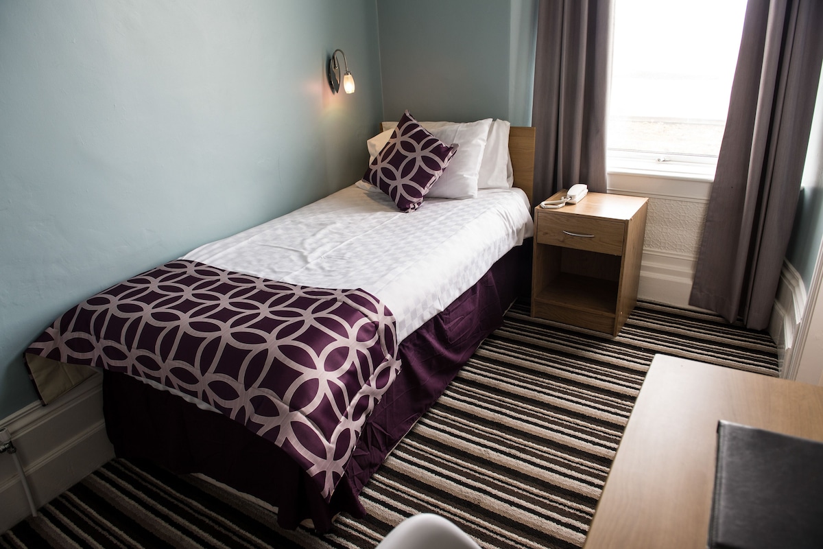 A single bed with a patterned bedspread, a bedside table with a lamp, and a window with curtains.