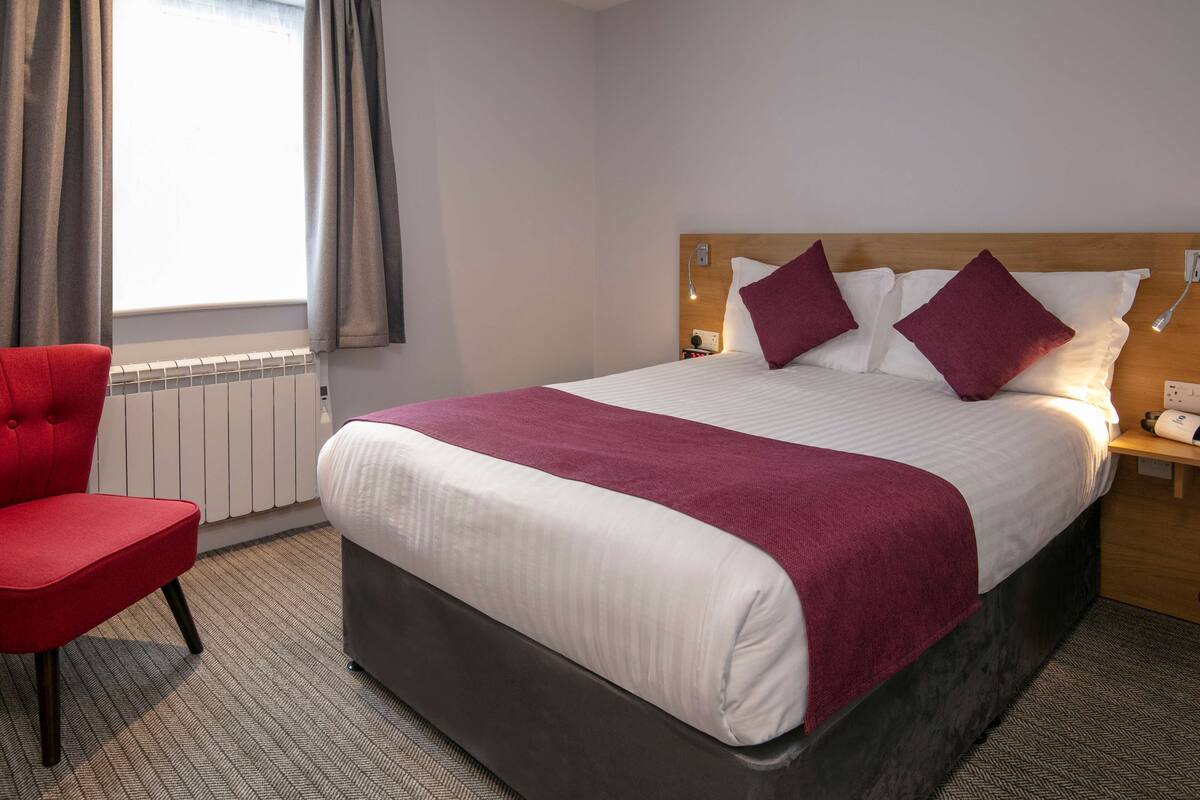 Una habitación de hotel con una cama grande, una silla roja, un cabecero de madera y una ventana con cortinas.
