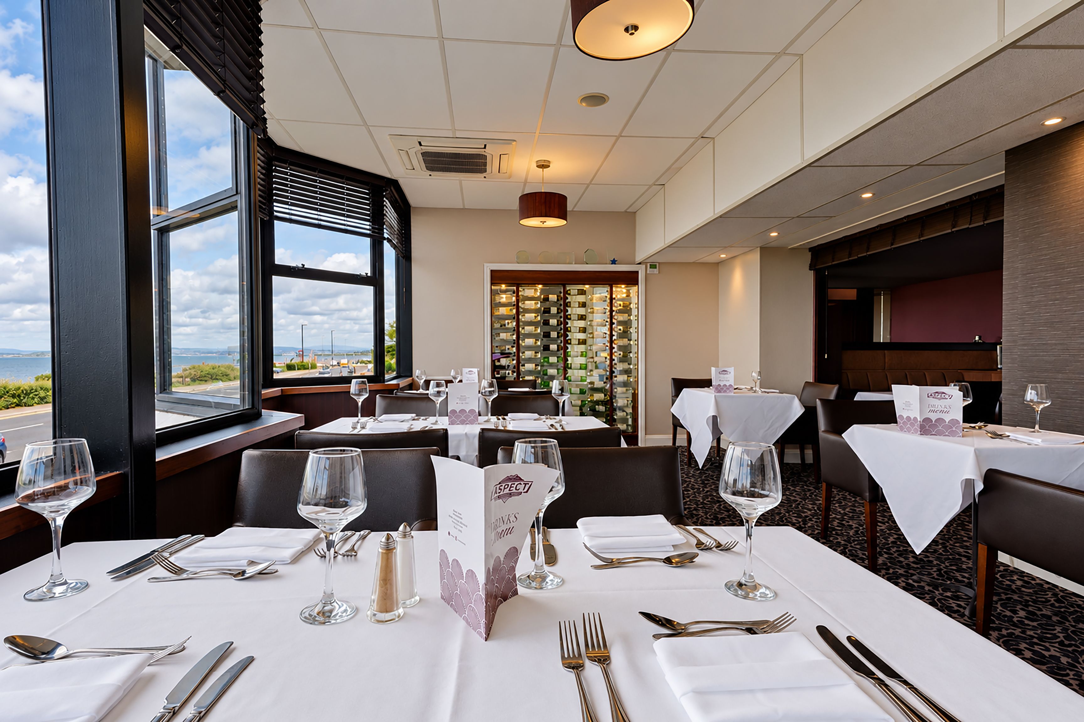 Ein Restaurant mit gedeckten Tischen, Meerblick durch große Fenster und einem Weinkühlschrank im Hintergrund.