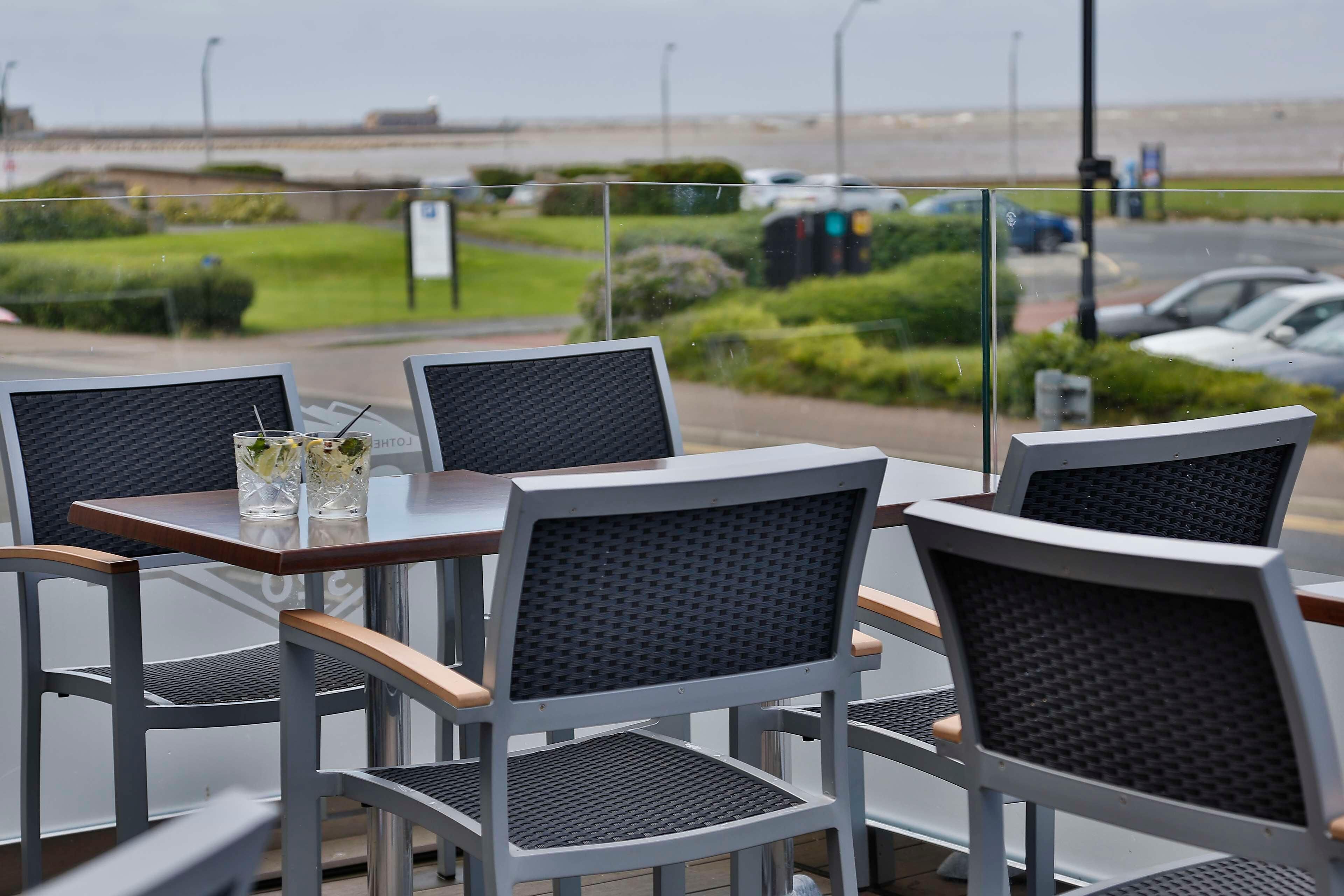 A table and chairs with a view of the sea and parked cars.