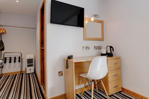 A hotel room with a wooden desk, a white chair, a flat-screen TV mounted on the wall, a wooden-framed mirror, and a coffee maker on the desk.