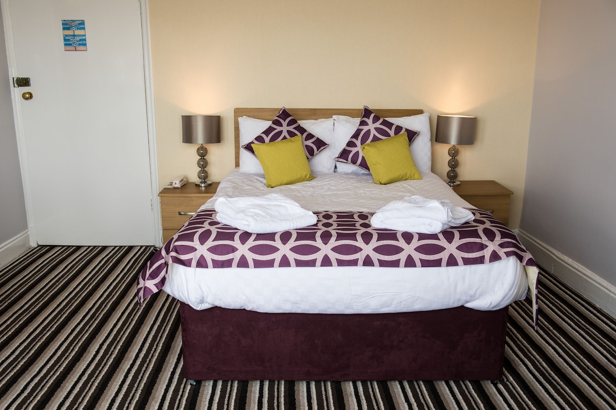 A neatly made bed with a patterned bedspread, two pillows, and two towels. There are two bedside lamps and a nightstand with a phone. The room has a striped carpet and a door with a lock.