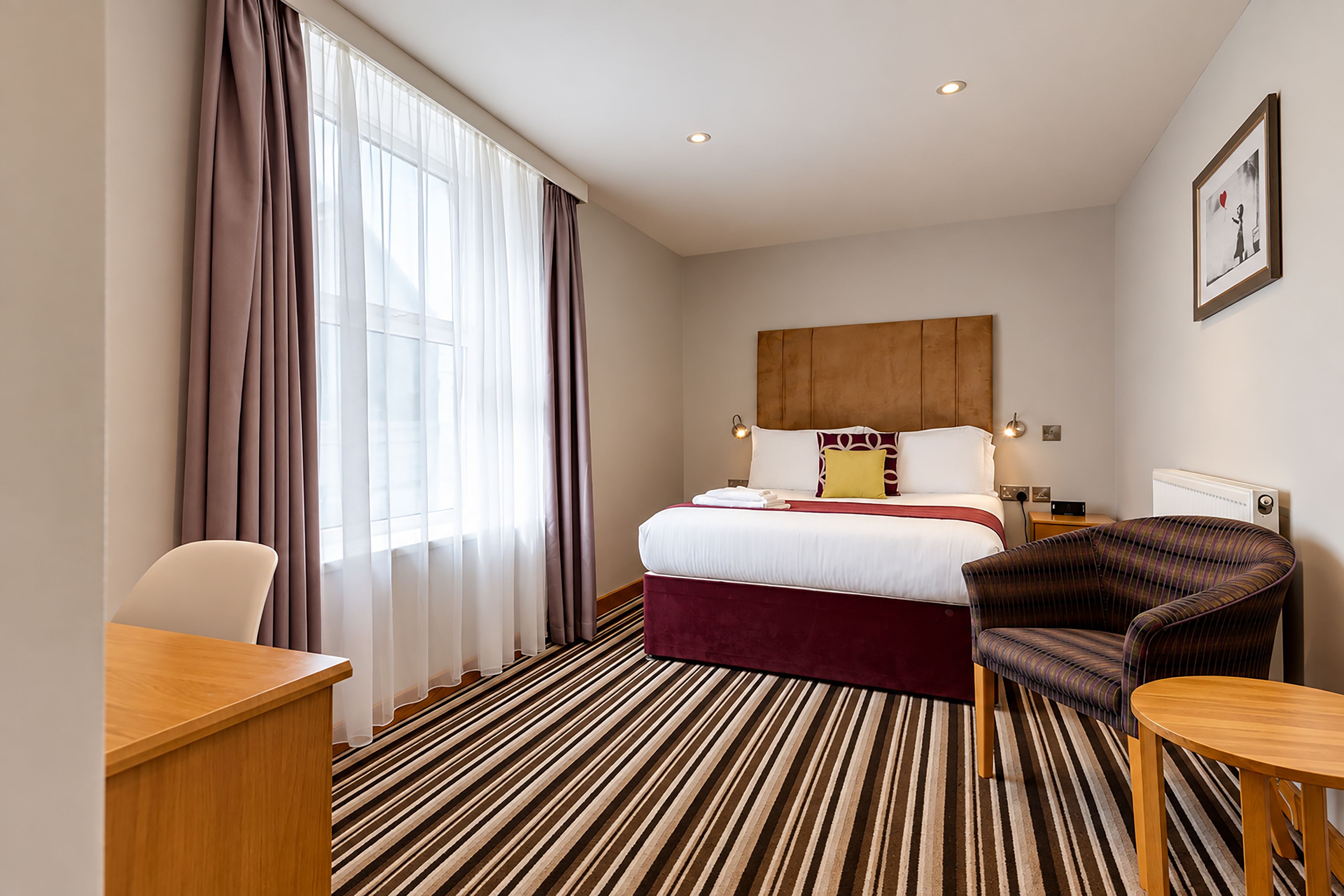 A hotel room with a bed, a desk, a chair, a radiator, and a window with curtains.