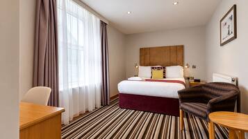 A hotel room with a bed, a desk, a chair, a radiator, and a window with curtains.