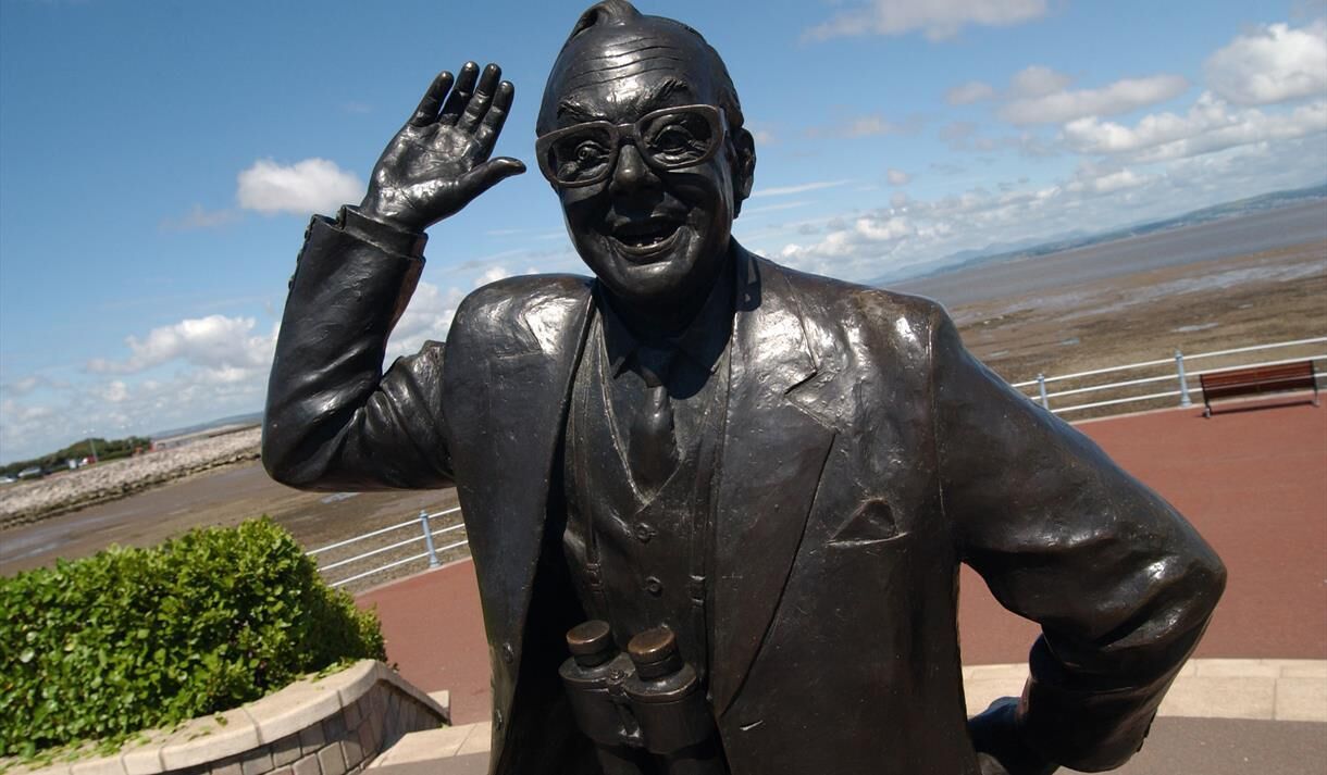 A statue of a man in a suit with a raised hand, located near a waterfront with a bench and railing.
