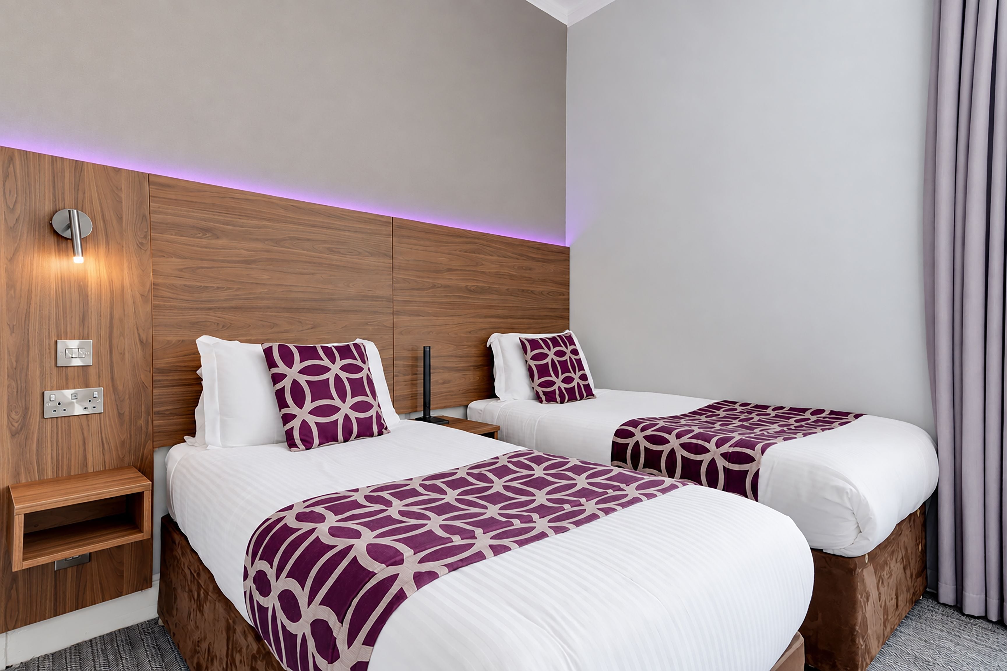 Habitación de hotel con dos camas, cabecero de madera, colchas estampadas color púrpura y una mesita de noche.