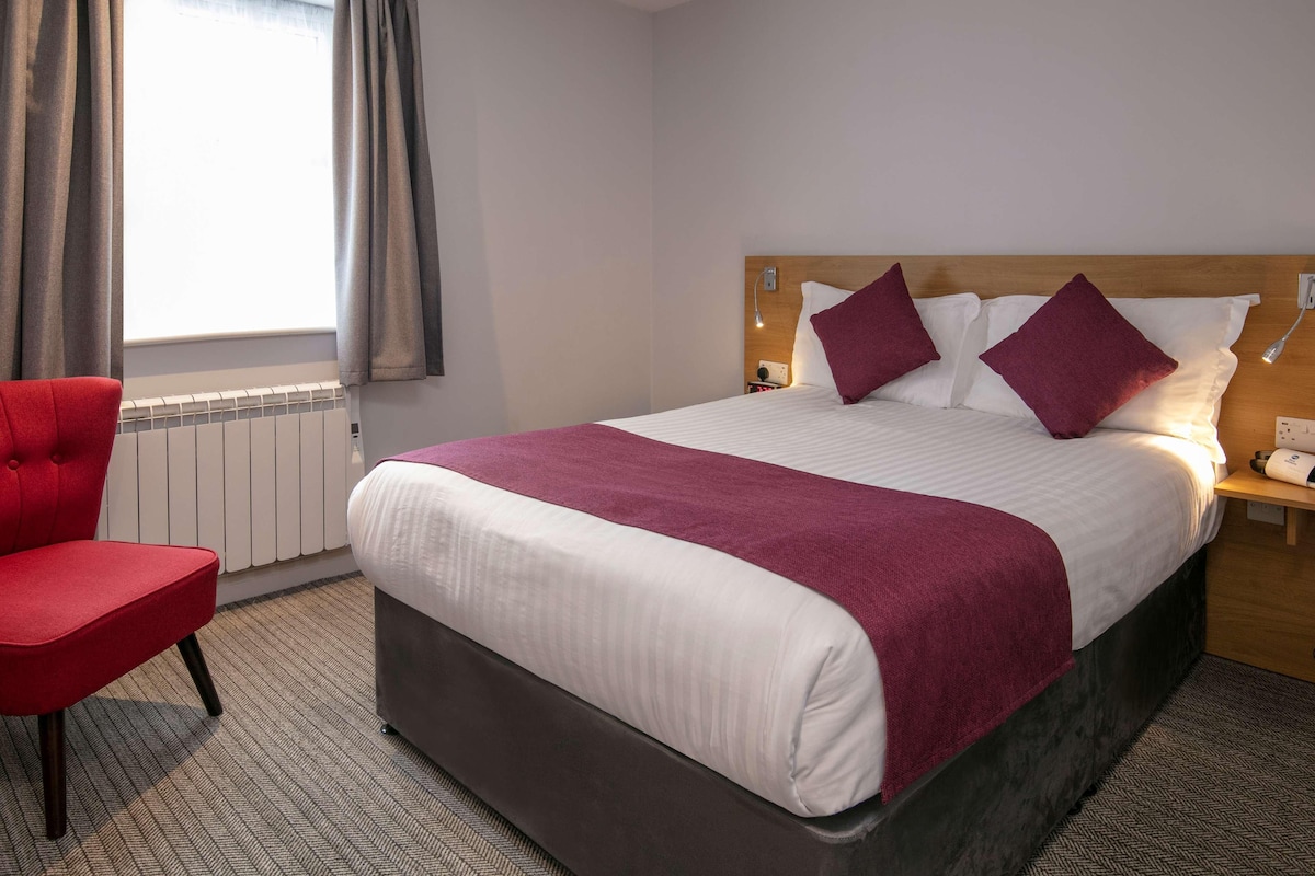 A hotel room with a bed, a red chair, a bedside table with a lamp, and a window with curtains.