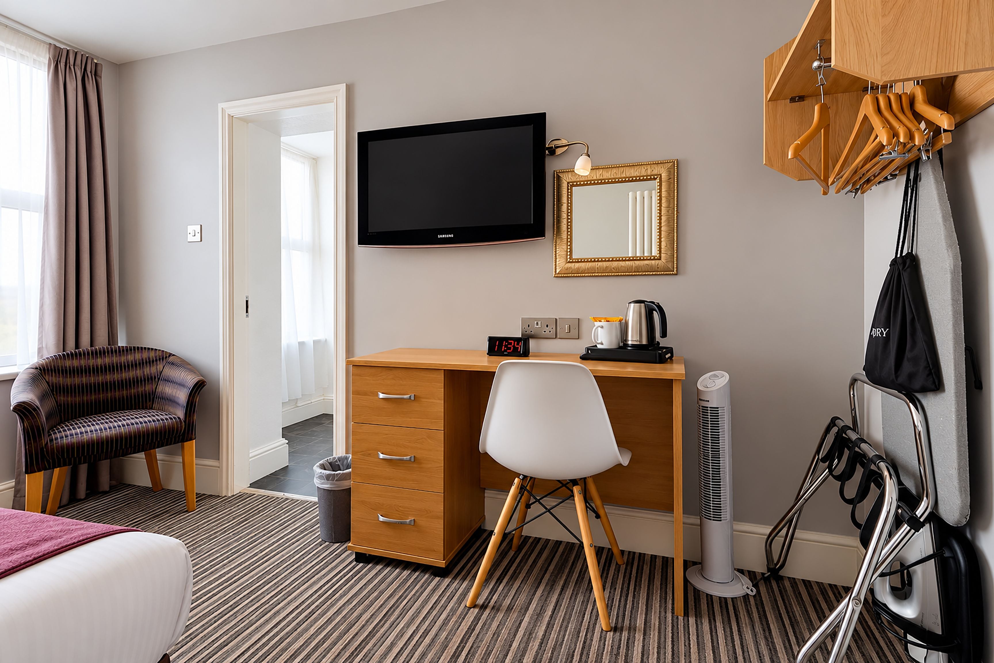 Hotelkamer met een bed, een bureau, een stoel, een televisie, een spiegel, een klok, een waterkoker, een ventilator en een kapstok.