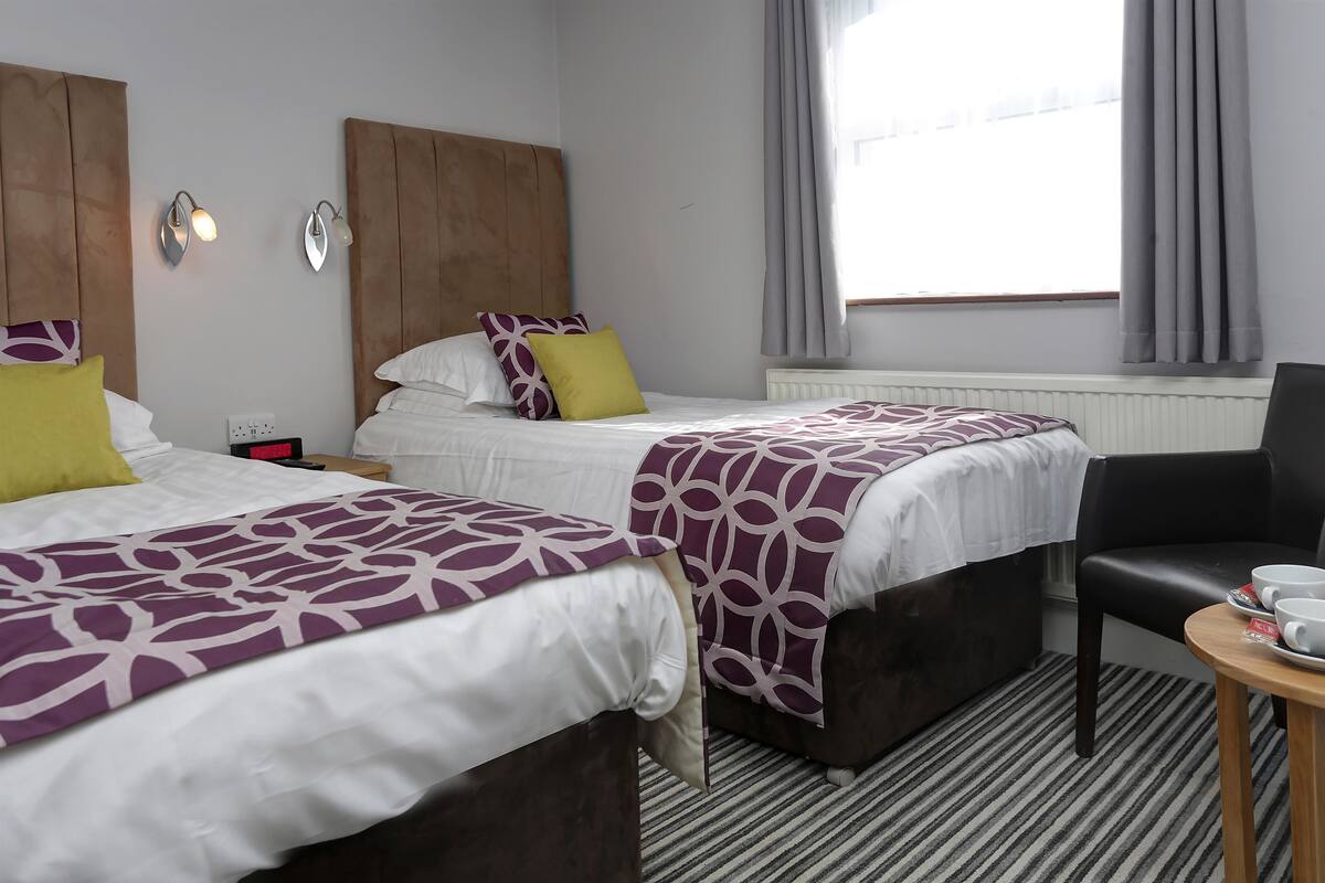 Habitación de hotel con dos camas, cada una con cabecera, mesita de noche, una silla y una pequeña mesa con tazas.
