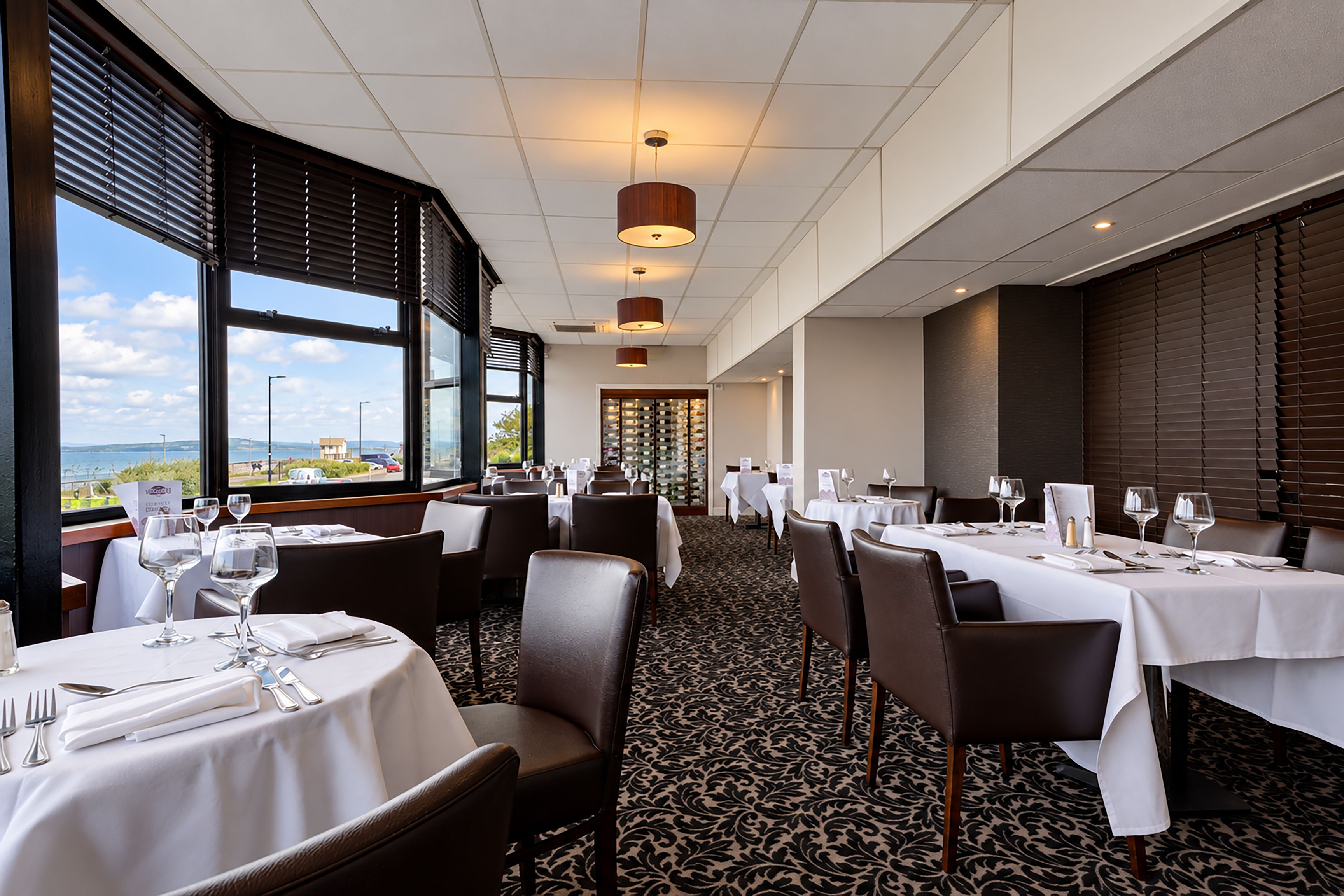 A restaurant with tables set for dining, large windows offering a view, and a patterned carpet.