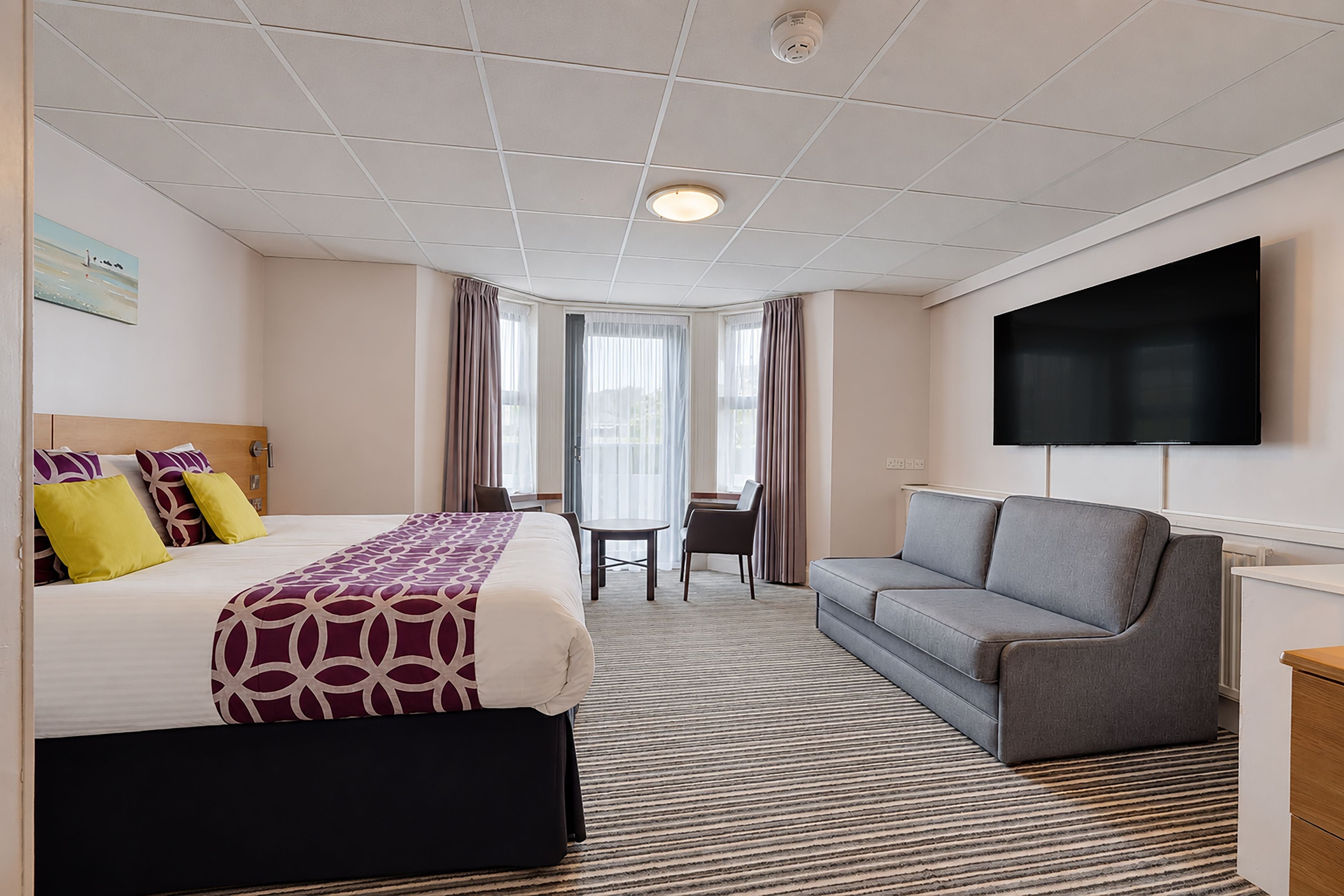 Ein Hotelzimmer mit Bett, einer Couch, einem Fernseher, einem Schreibtisch und einem fensterbepflanzten Fenster.