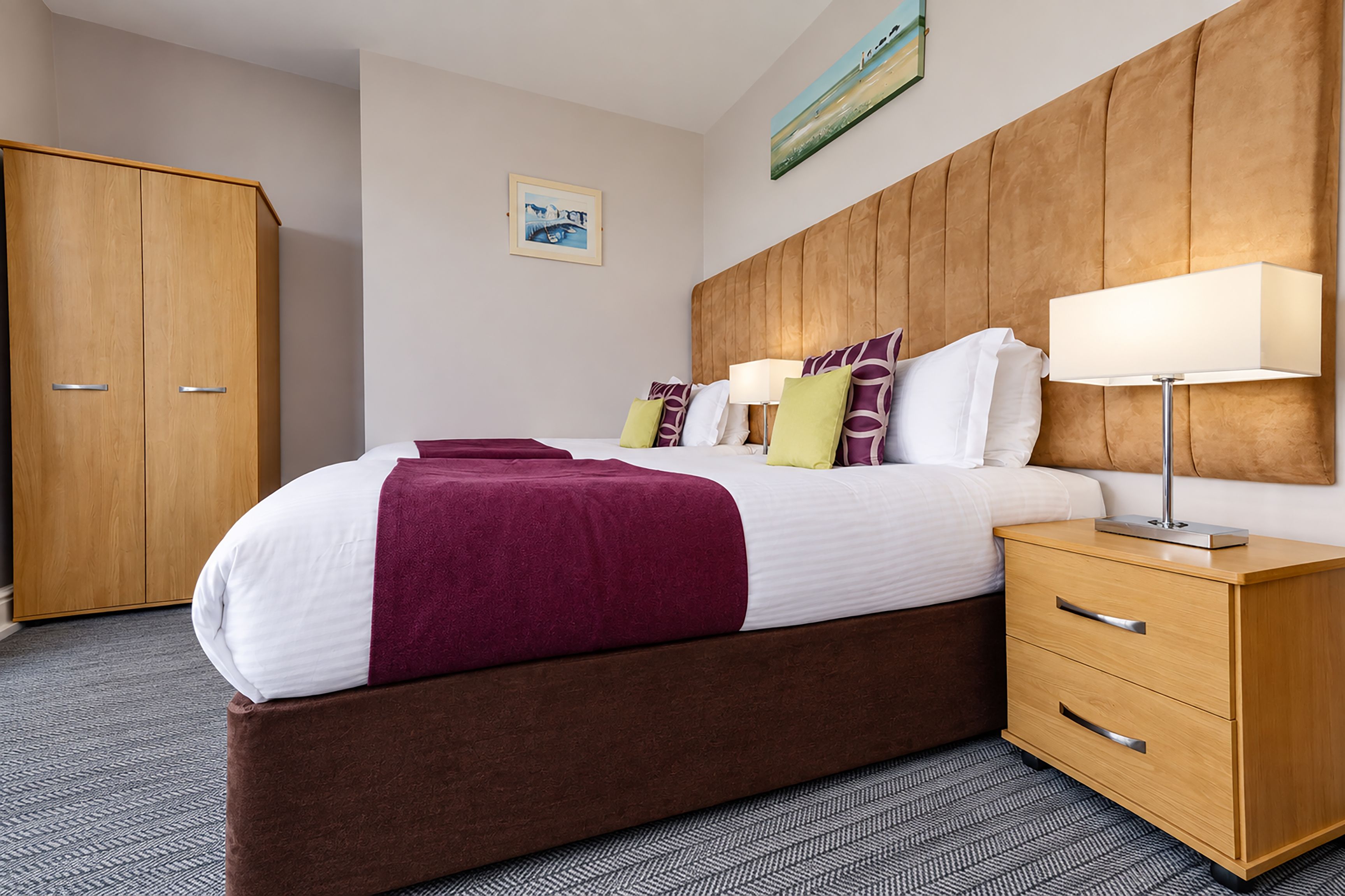 A hotel room with a large bed, wooden headboard, a nightstand with a lamp, a wardrobe, and a painting on the wall.