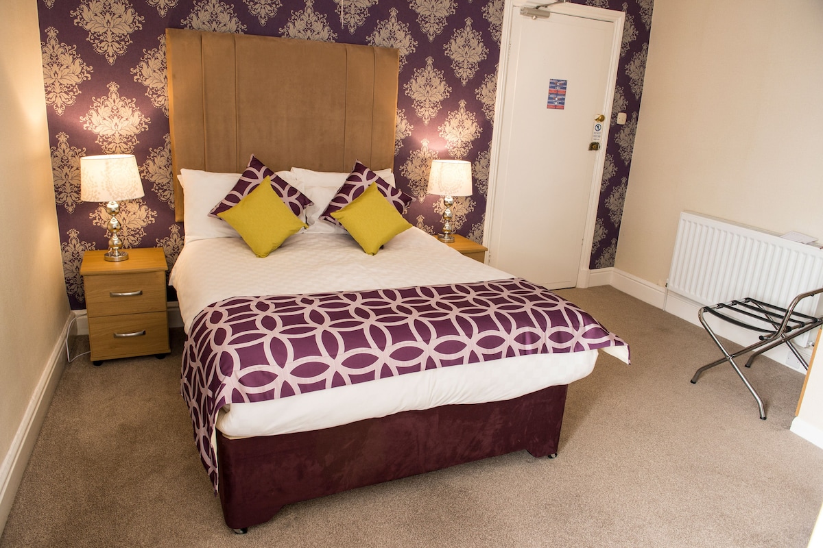 A neatly made bed with a patterned bedspread and pillows, two bedside tables with lamps, a wooden nightstand with a drawer, a radiator, and a wall with a floral wallpaper pattern.