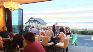 Restaurante con terraza al aire libre que da a una masa de agua, con candelabro y vista a una casa en el fondo.