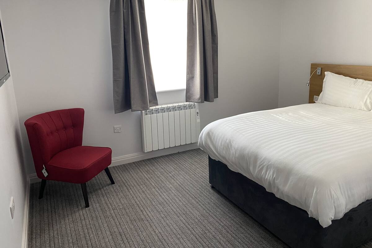 Una habitación de hotel con una cama, una silla roja, un radiador y cortinas grises.