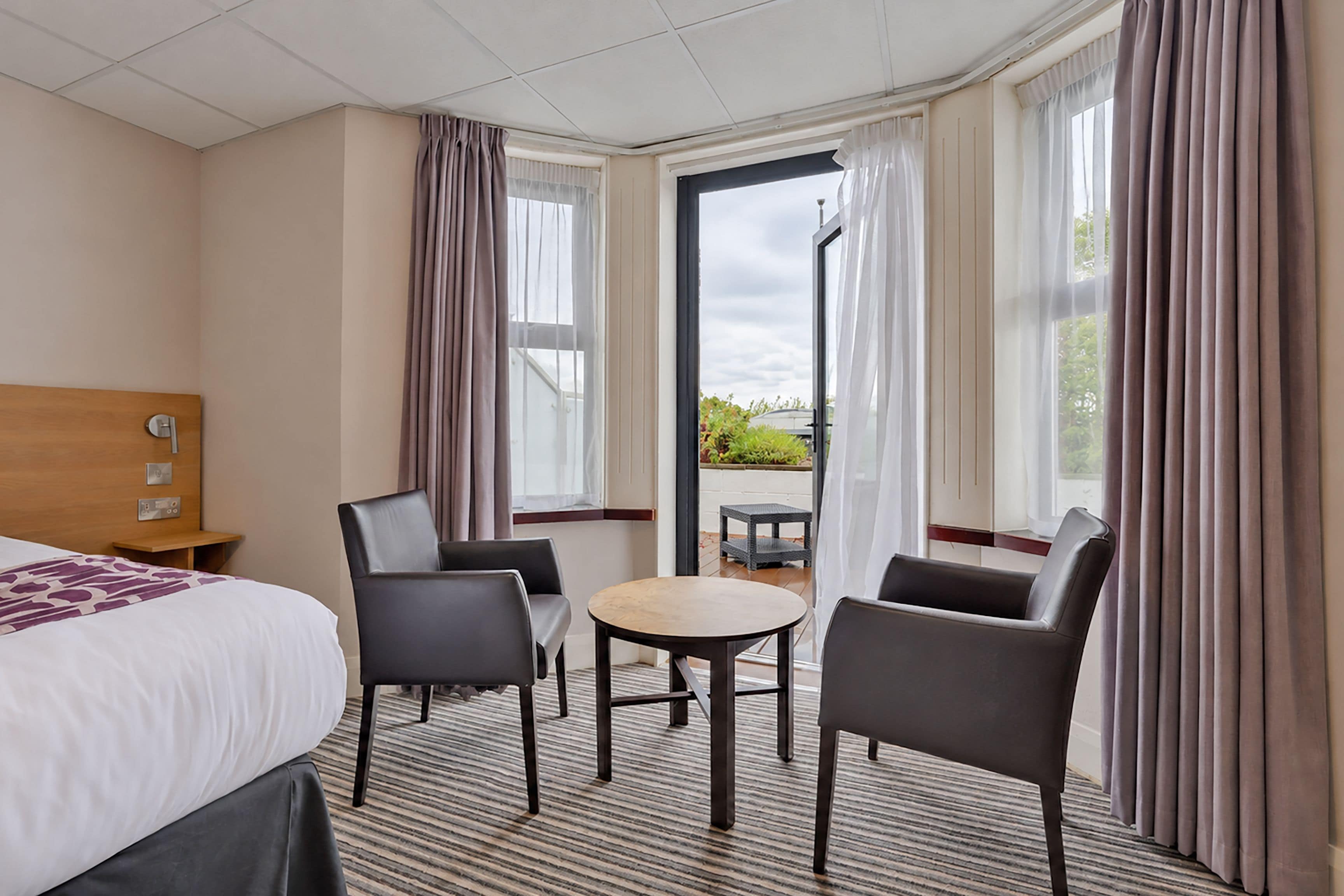 A hotel room with a bed, two chairs, a small table, and a view of greenery through the window.