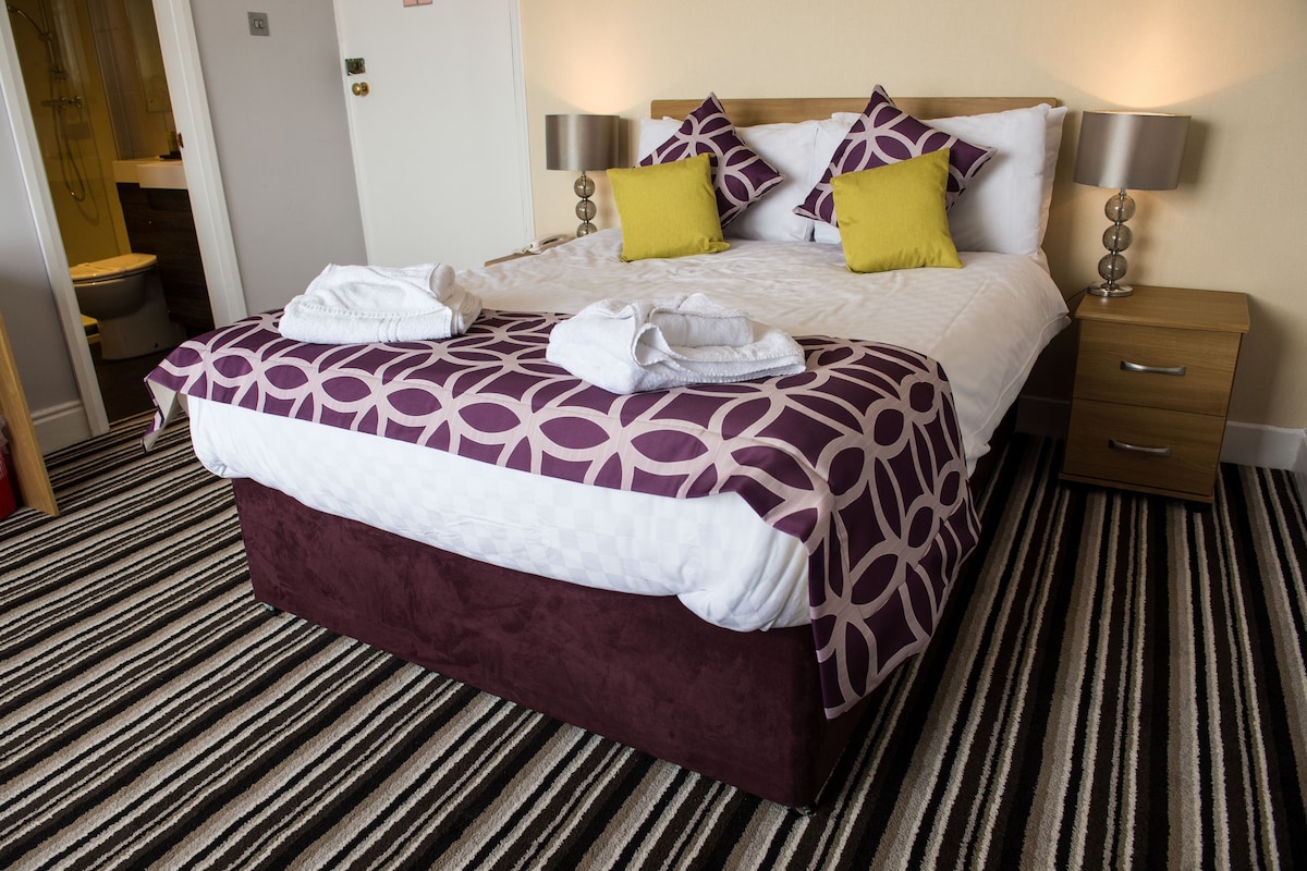 A hotel room with a large bed, two bedside tables, a wooden nightstand, and a bathroom visible through an open door.