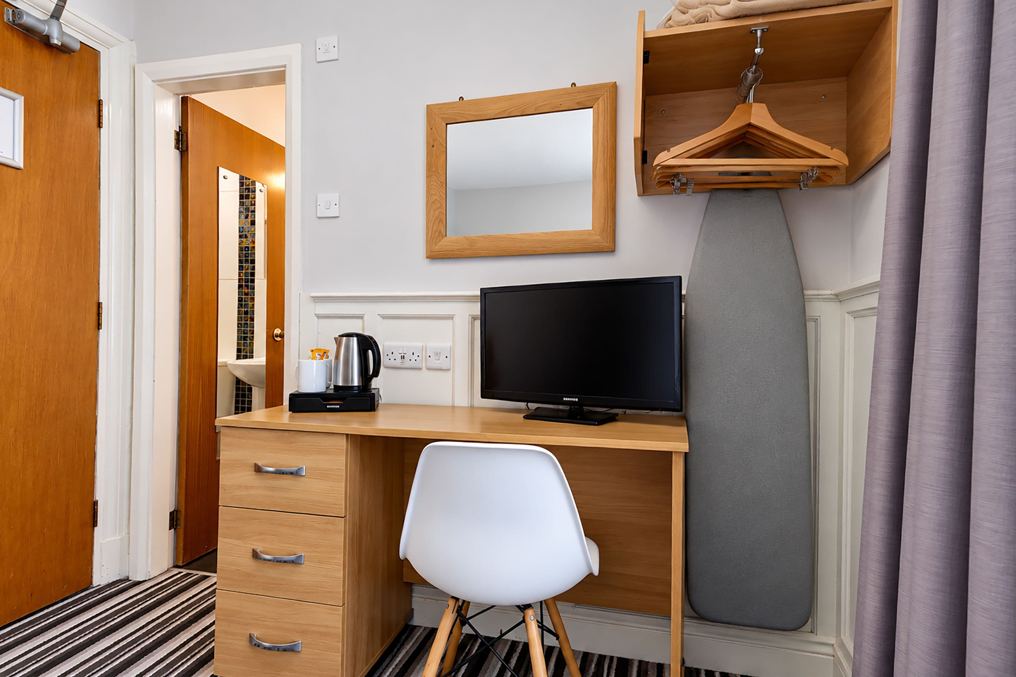 Habitación de hotel con escritorio de madera, televisión de pantalla plana, silla blanca, perchero y tabla de planchar gris.