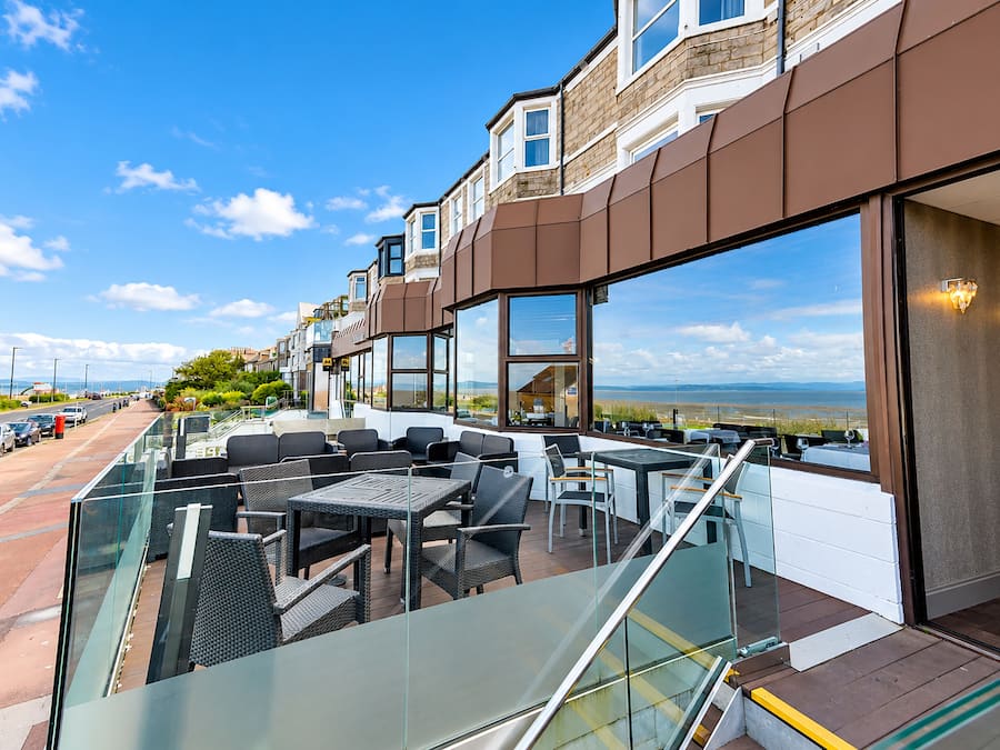 Terraço com mesas ao ar livre, vista para o mar e um edifício com grandes janelas.