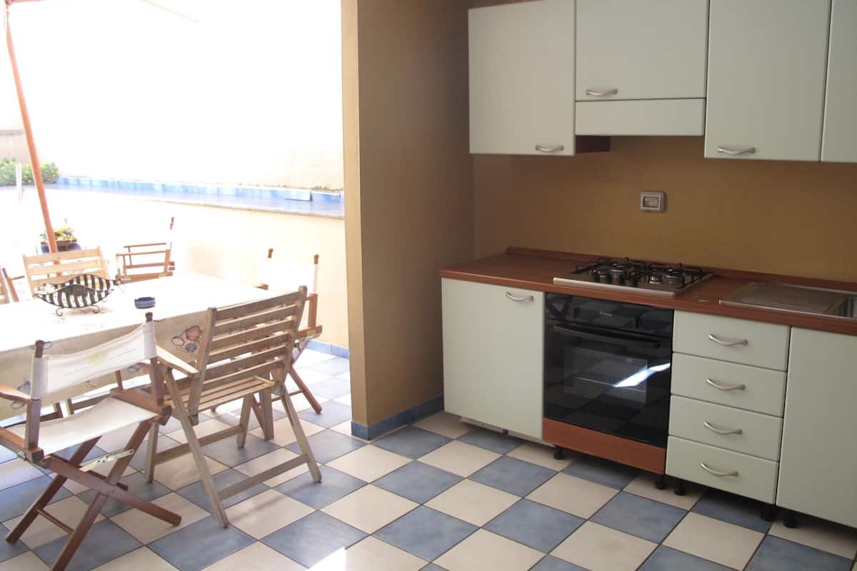 A compact kitchen with a gas stove, oven, and white cabinets. There is a dining table with chairs and a small table with a decorative item. The floor has a checkered tile pattern.