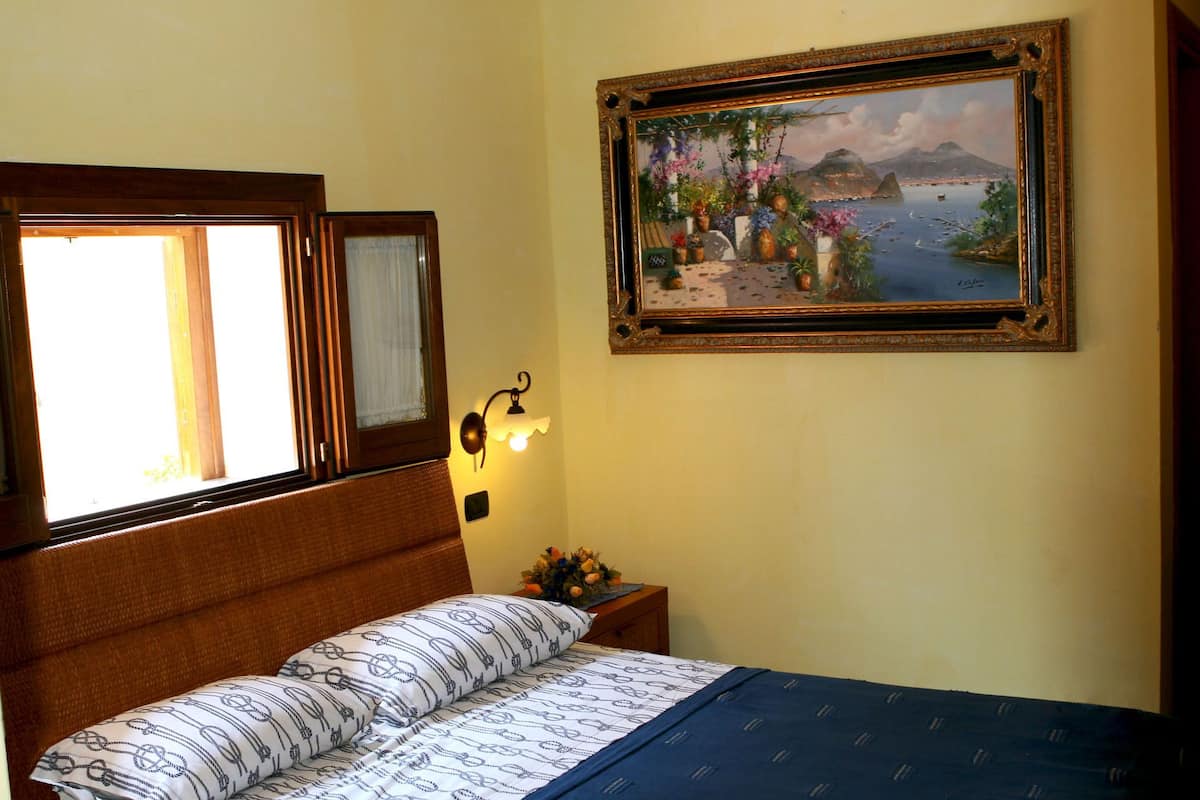 A bedroom with a bed, a nightstand with a lamp, a window with curtains, and a painting on the wall.