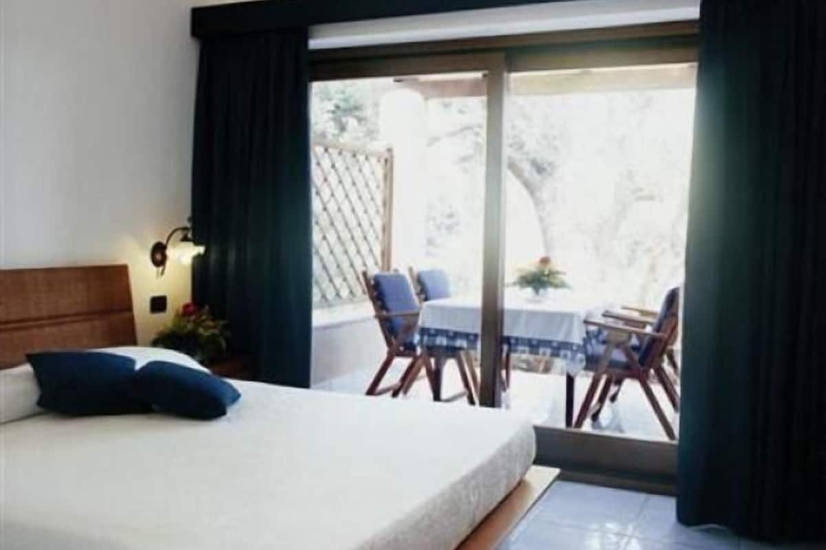 A bedroom with a bed, a nightstand, a window with curtains, and a view of an outdoor patio area with a table and chairs.