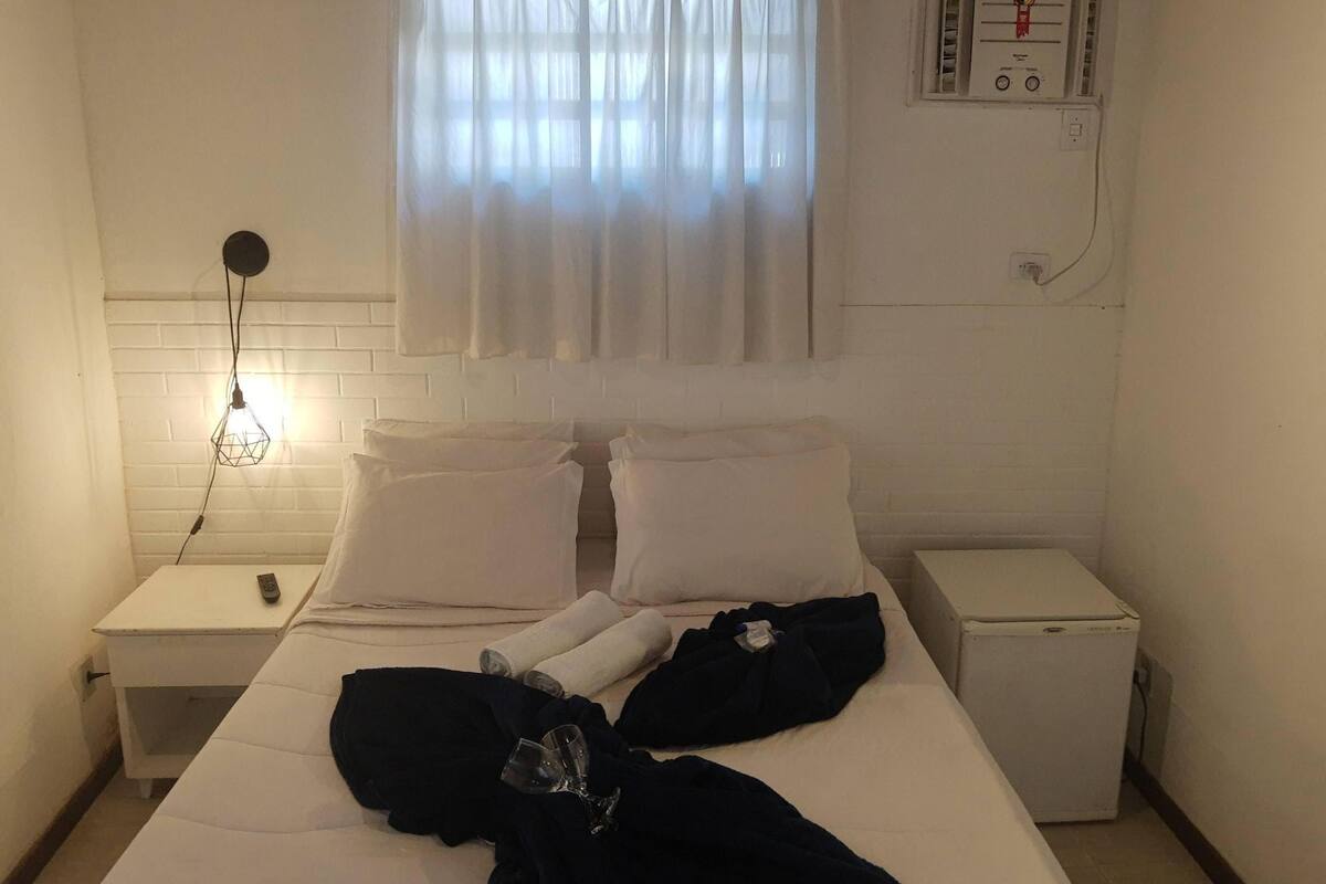 Habitación de hotel con cama, mesita de noche, un calentador pequeño, ventana con cortinas y un aire acondicionado instalado en la pared.