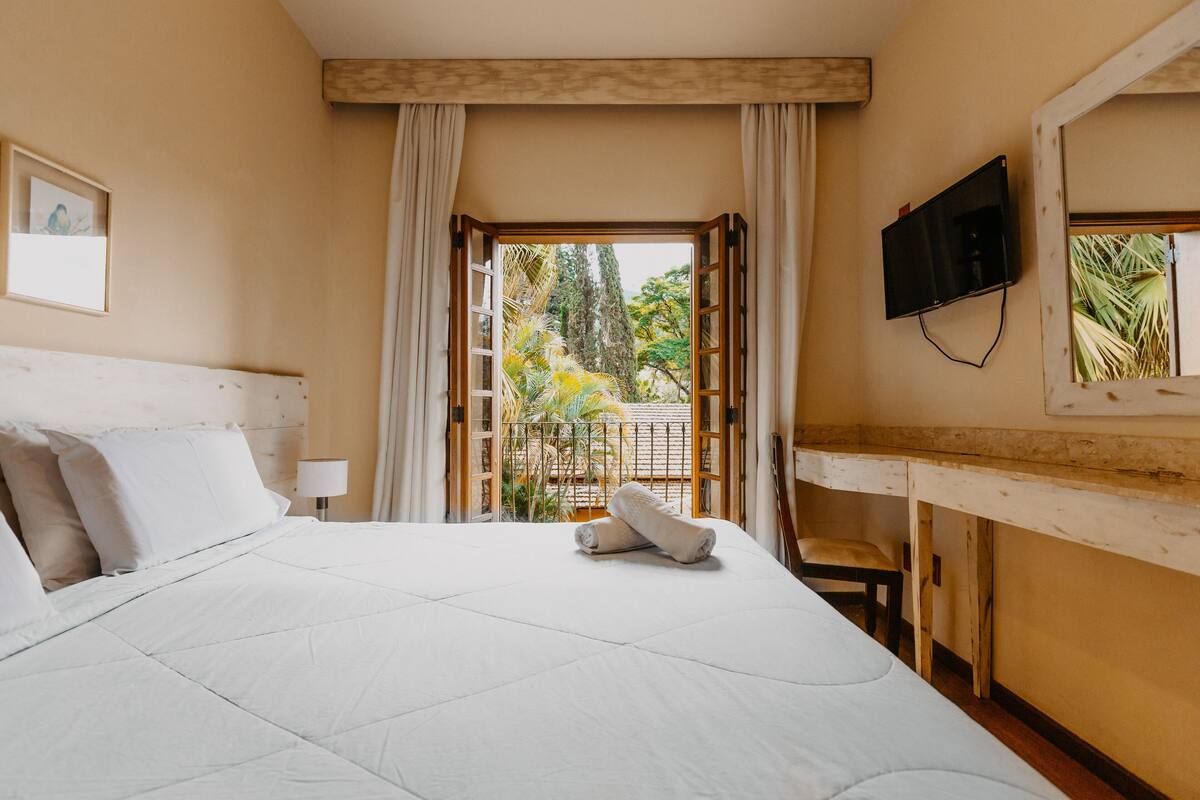 Una habitación de hotel con cama, televisor montado en la pared y balcón con cortinas.