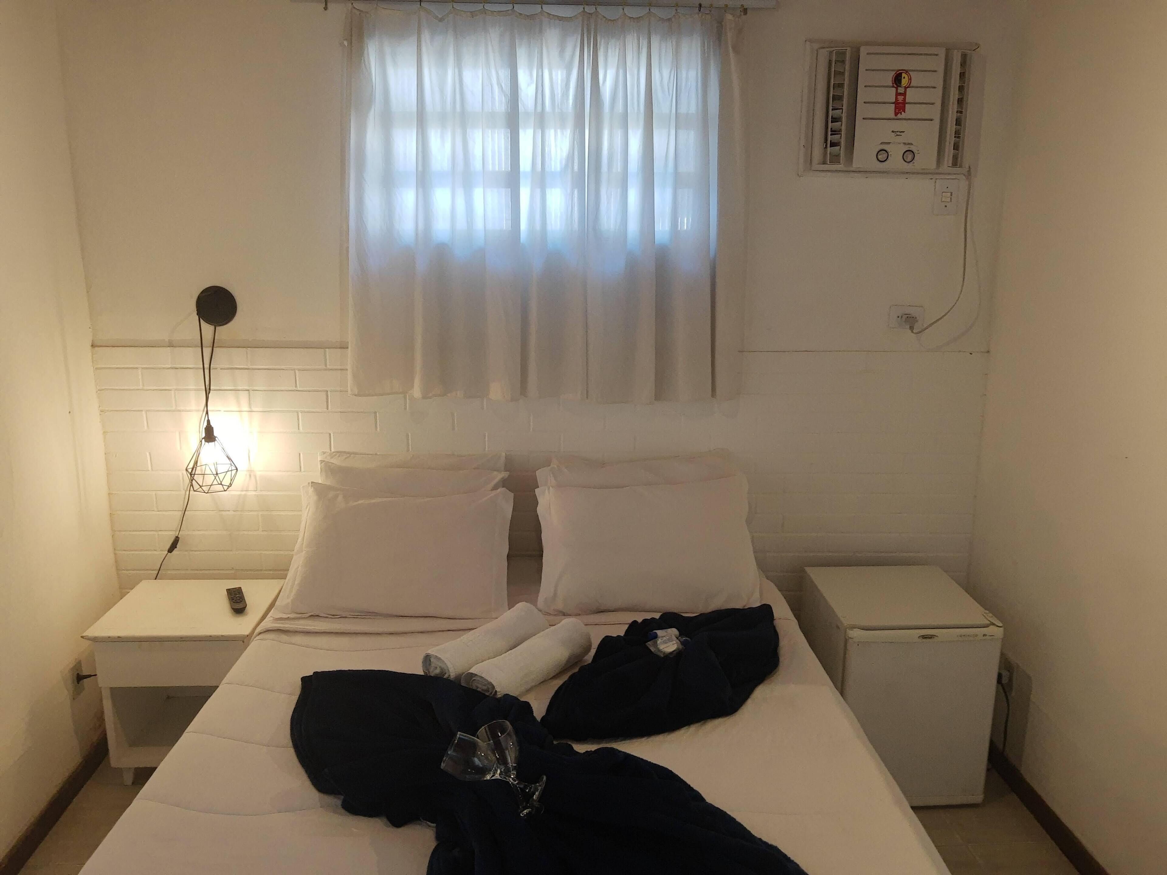 Quarto de hotel com uma cama, mesa de cabeceira, pequena mesa, aquecedor e janela com cortinas.