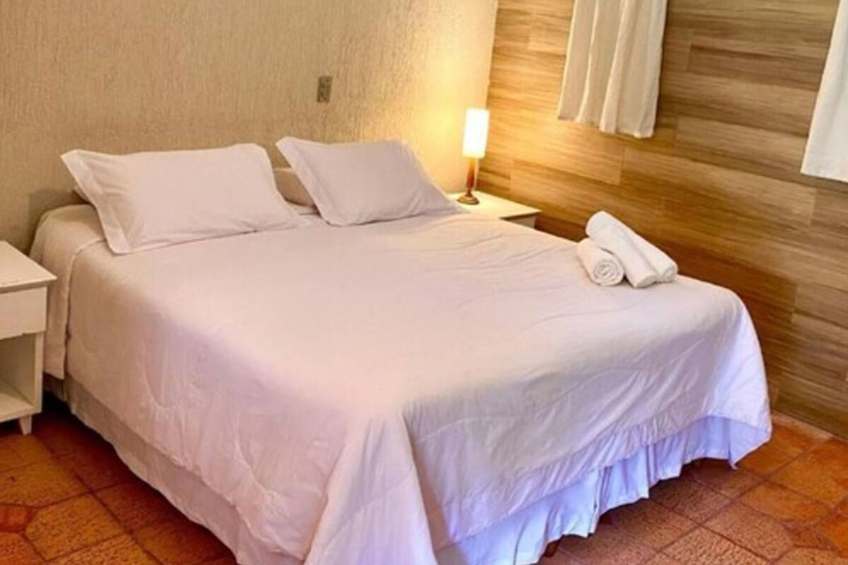 Una cama bien hecha con sábanas blancas, una lámpara de noche y una toalla enrollada sobre la cama.