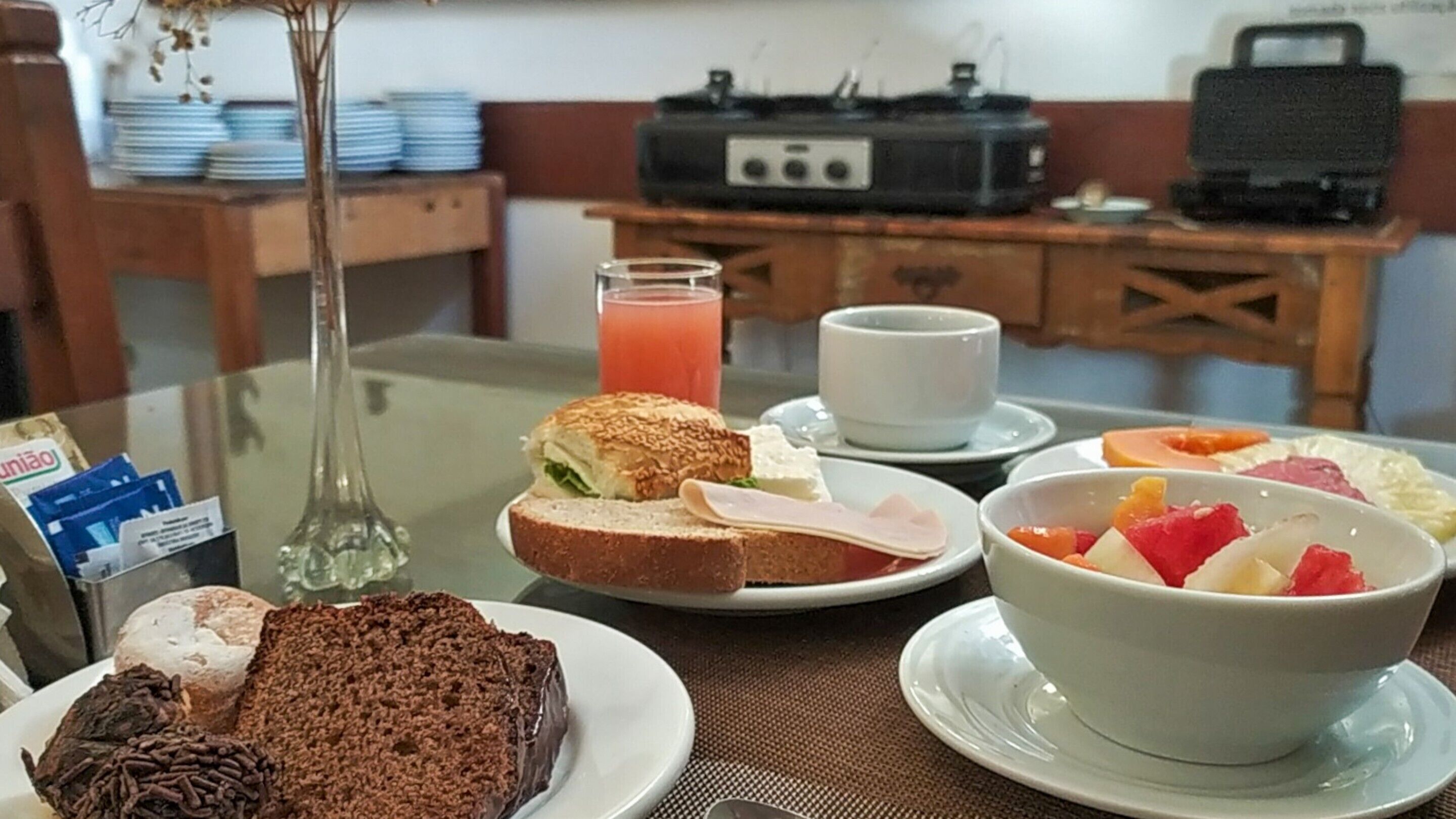 A breakfast setting with a variety of food items, including a slice of cake, a sandwich, a bowl of fruit, and a glass of juice.
