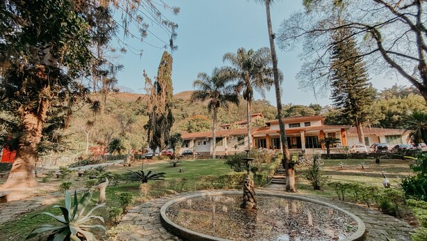 A landscaped garden with a circular pond, palm trees, and a building with a tiled roof in the background. - Hotel Vila Bavaria (Petrópolis)