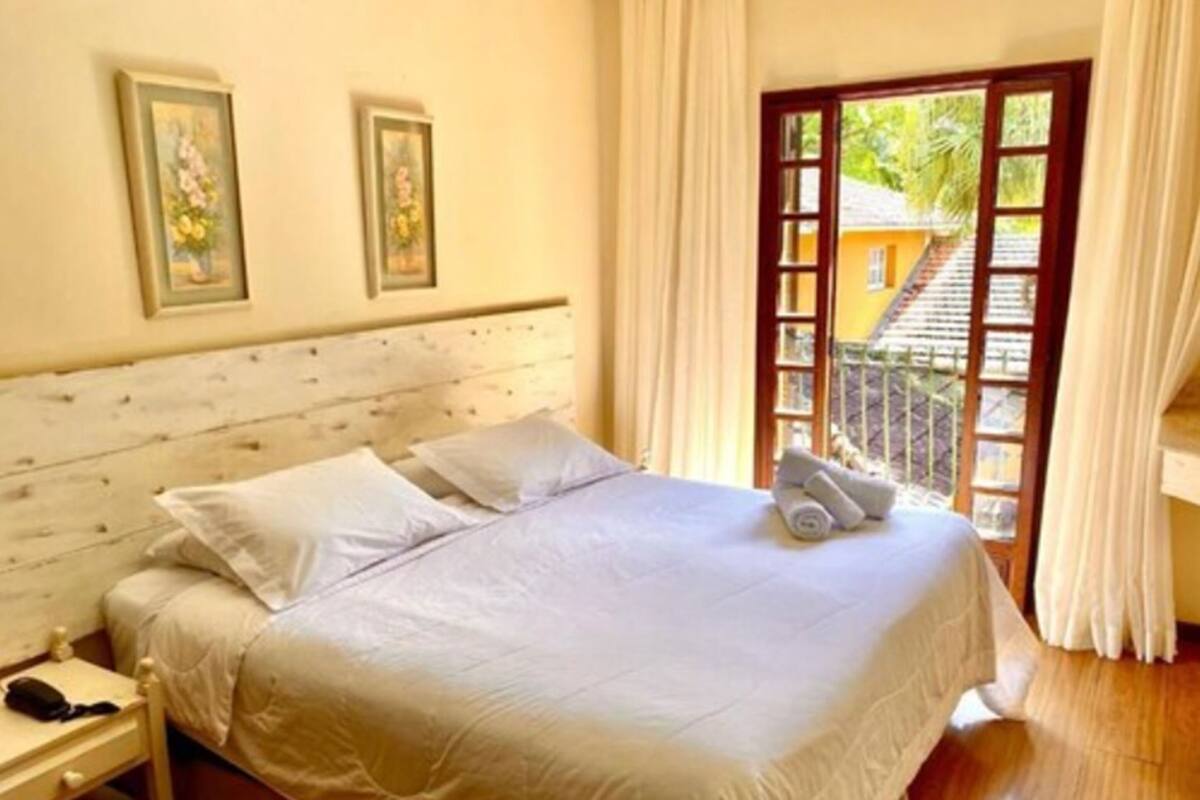 Un dormitorio con cabecera de madera, una cama con ropa de cama blanca, dos cuadros en la pared y vistas a un balcón a través de una puerta de cristal.