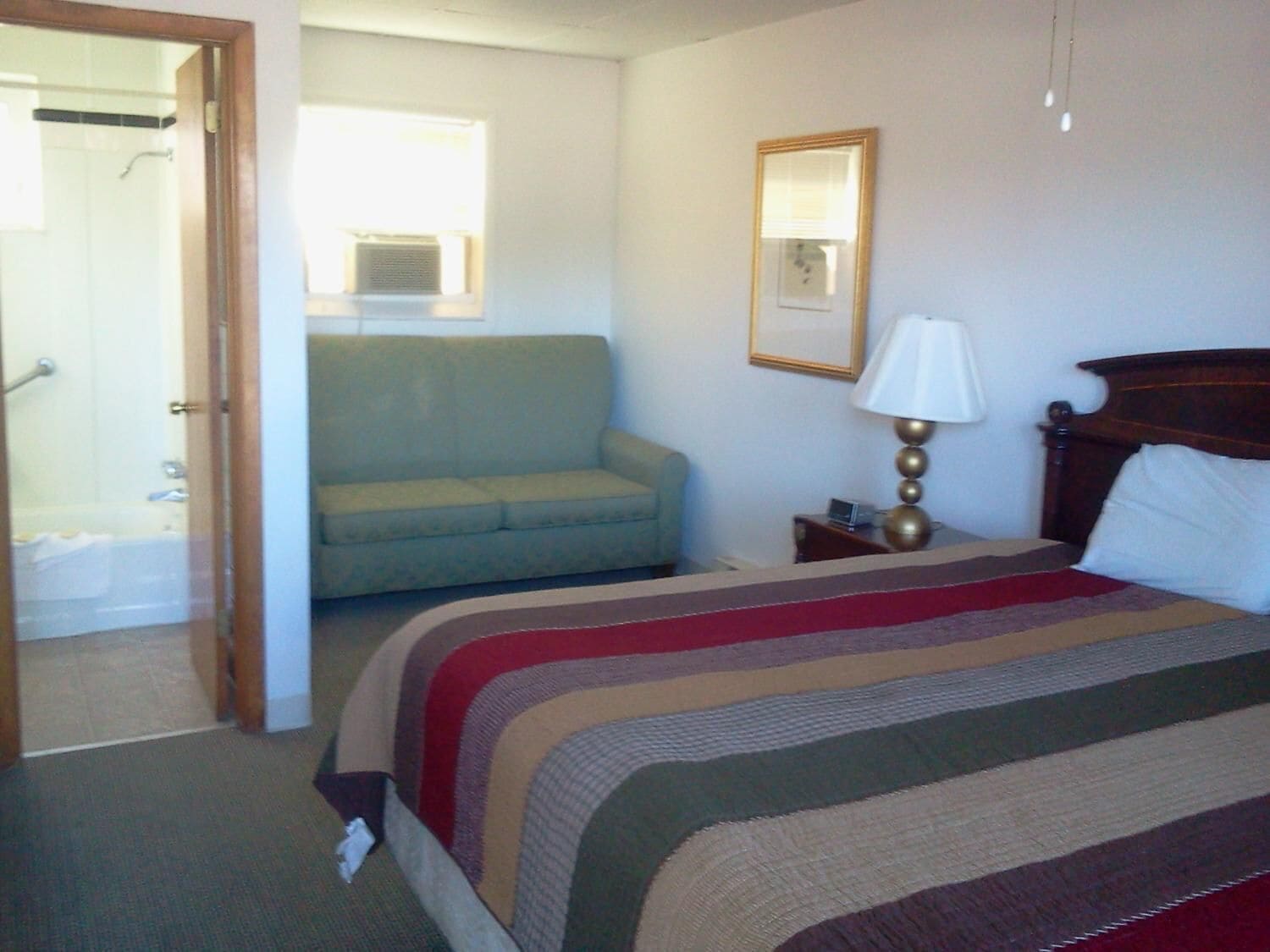 Una habitación de hotel con una cama, un sofá, una mesita de noche con lámpara, una ventana y una puerta que da a un baño.