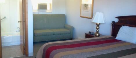 A hotel room with a bed, a sofa, a bedside table with a lamp, a window, and a door leading to a bathroom.