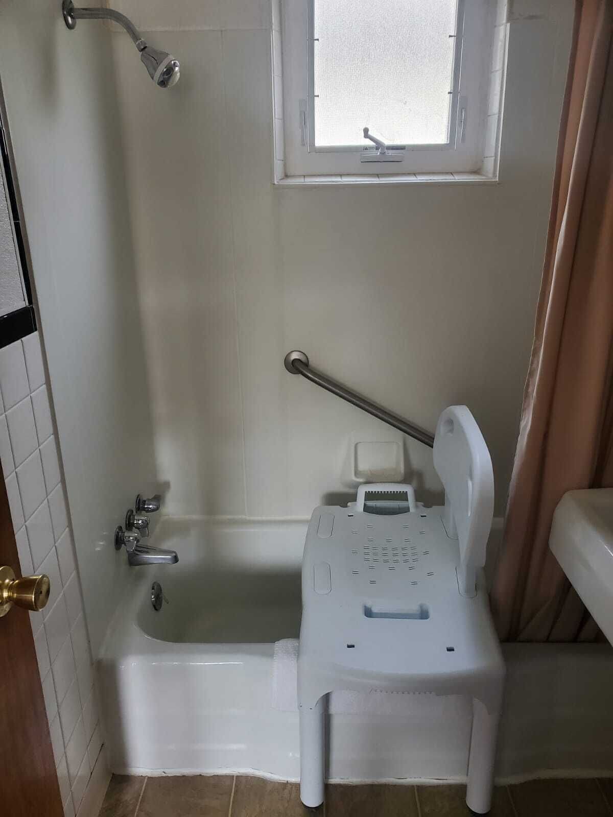 A bathroom with a bathtub, a window, a showerhead, and a small white chair.