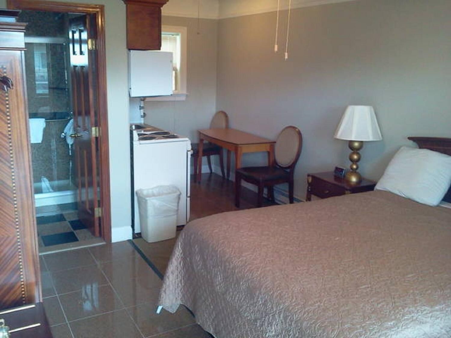 A hotel room with a bed, a desk, a chair, a lamp, and a small kitchenette.