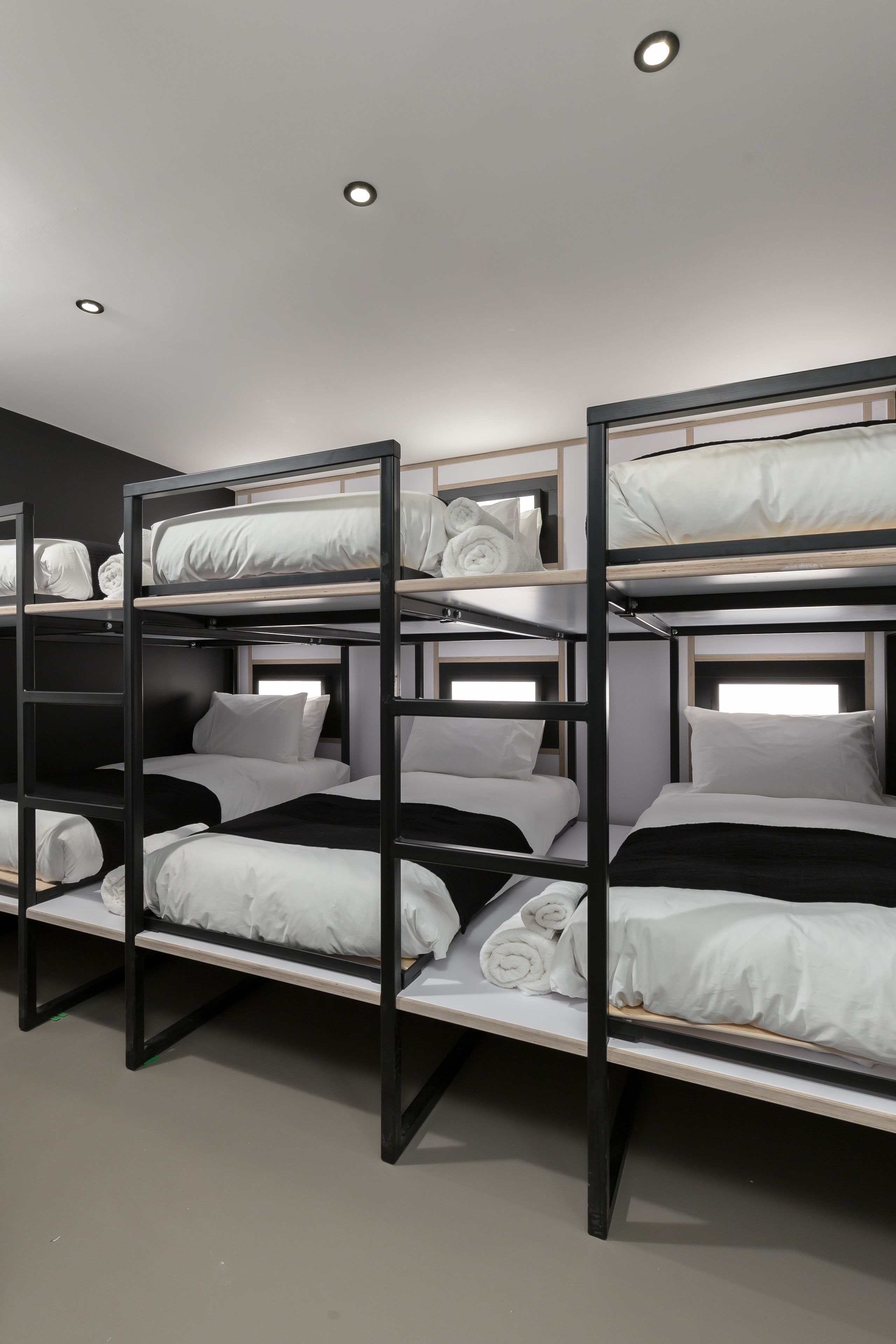 A bunk bed room with multiple beds, each with white bedding and pillows, in a modern room with a dark wall and ceiling lights.