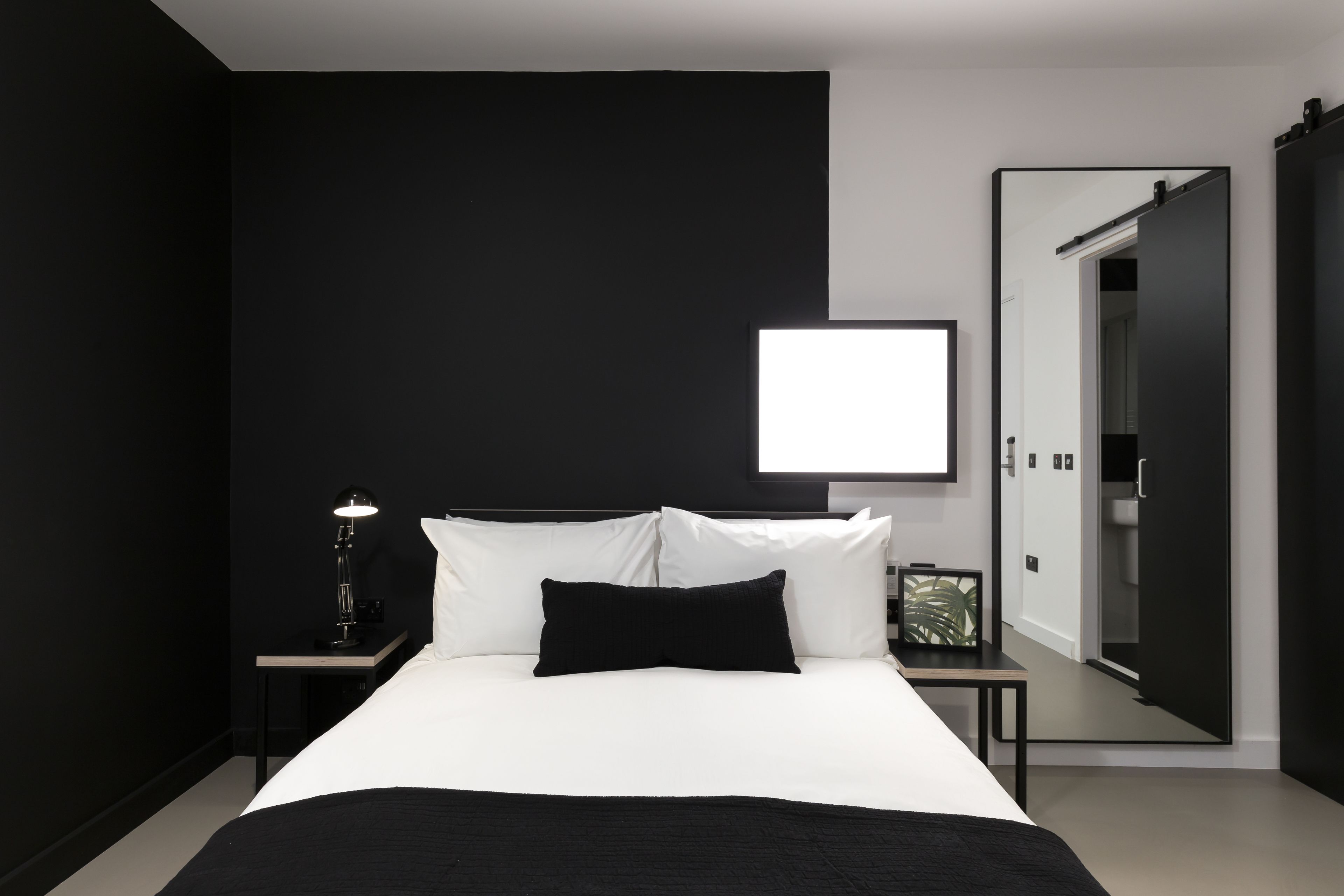 A modern bedroom with a large bed, a bedside table with a lamp, a framed picture, and a mirror on the wall.